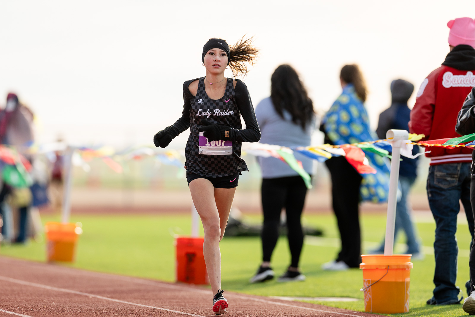 3A-4A-district-xc-jpegJoe-pps-17 | Press Pass Sports Female runner in a black checkered jersey labeled'Lady Raiders' sprinting on a track with colorful pennant banners in the background.
