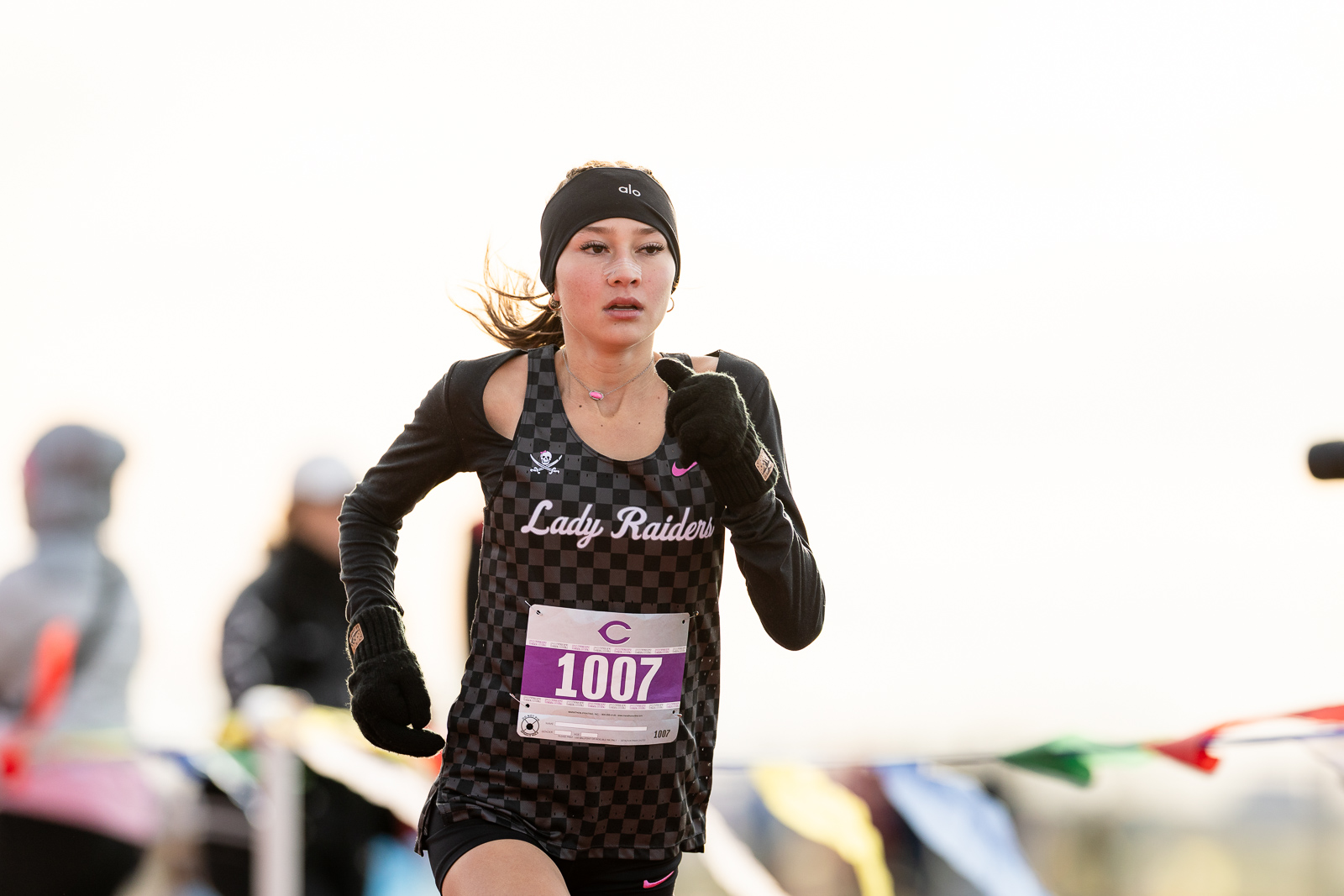 3A-4A-district-xc-jpegJoe-pps-18 | Press Pass Sports Female runner in a checkered black-and-gray racing jersey with'Lady Raiders' on the chest, racing bib 1007, headband, black gloves, mid-stride outdoors.