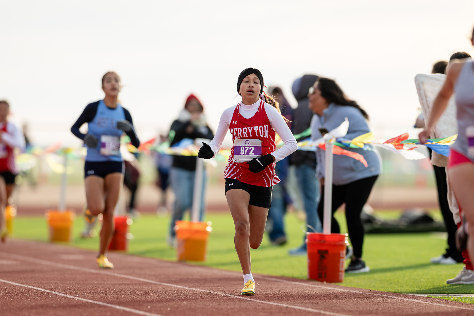 3A-4A-district-xc-jpegJoe-pps-19 | Press Pass Sports Young female runner in red uniform with bib 972 leading a track race on an outdoor field, spectators cheering in the background.