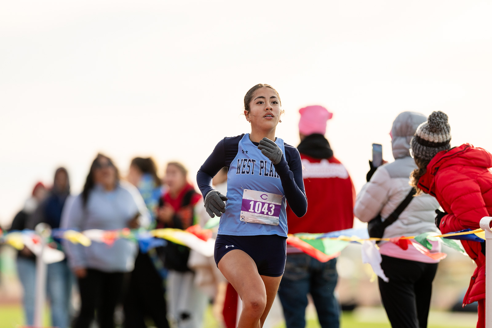 3A-4A-district-xc-jpegJoe-pps-20 | Press Pass Sports Female runner with bib 1043 in a light blue West Plains singlet, sprinting toward the finish line as spectators cheer behind colorful tape.