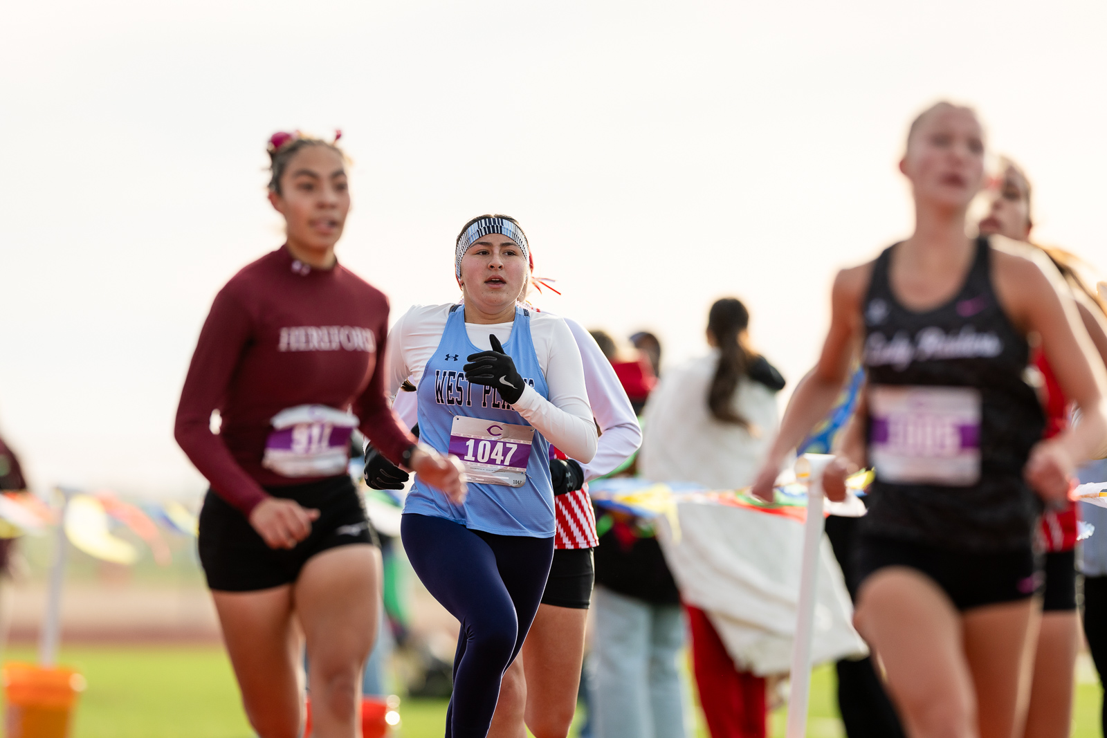 3A-4A-district-xc-jpegJoe-pps-21 | Press Pass Sports Central runner in a light blue top with white sleeves and striped headband, bib 1047, mid-race with other runners blurred in the background.