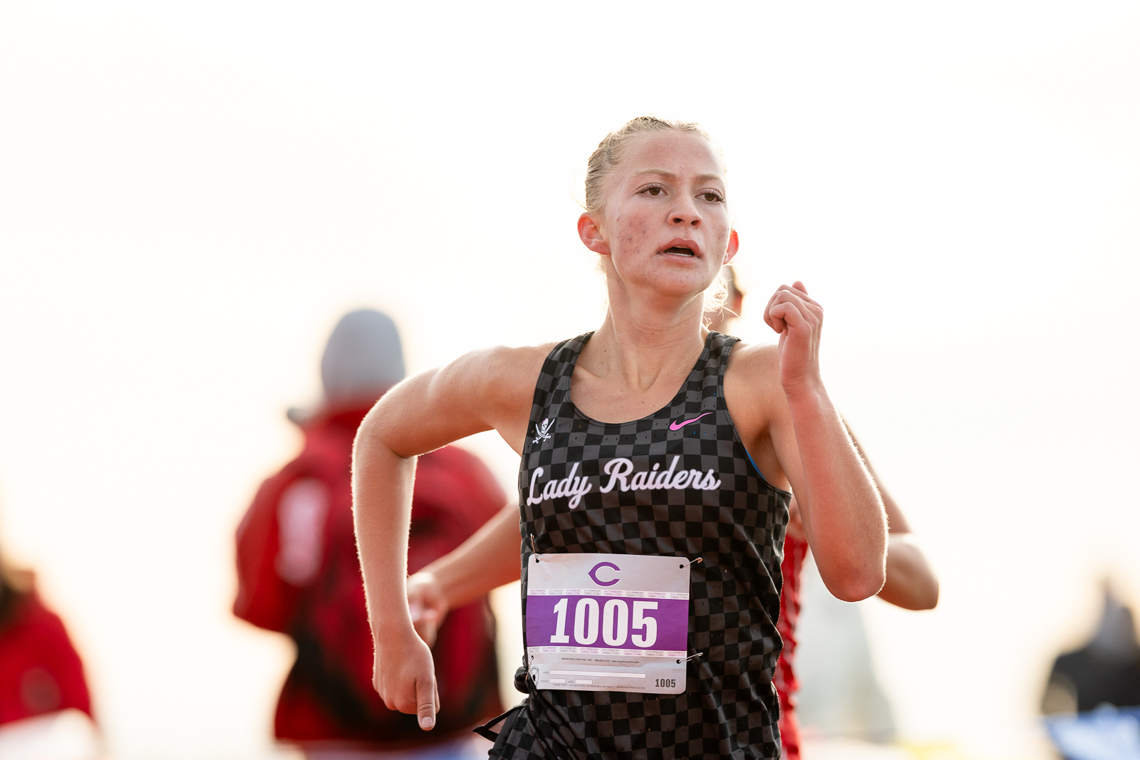 3A-4A-district-xc-jpegJoe-pps-22 | Press Pass Sports Young female runner in a black checkered tank top reading'Lady Raiders' with race bib 1005, mid-stride in a race.
