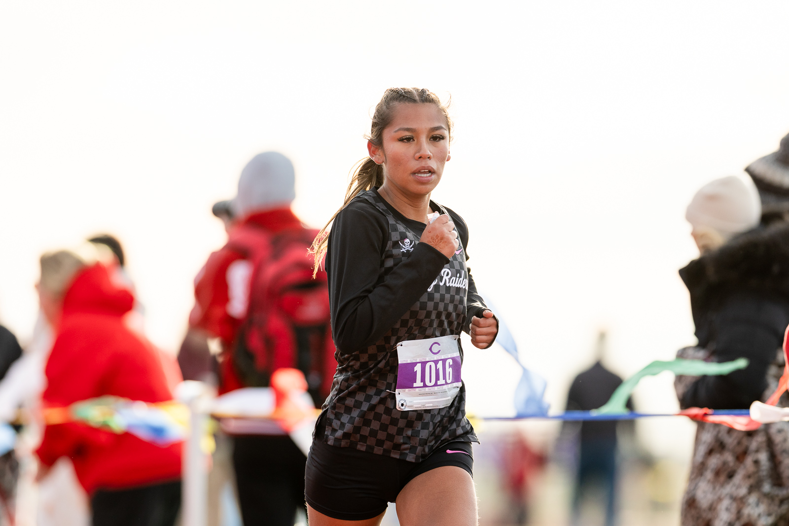 3A-4A-district-xc-jpegJoe-pps-25 | Press Pass Sports Female runner in a black checkered jersey with bib 1016 races forward, focused expression, spectators in the blurred background.