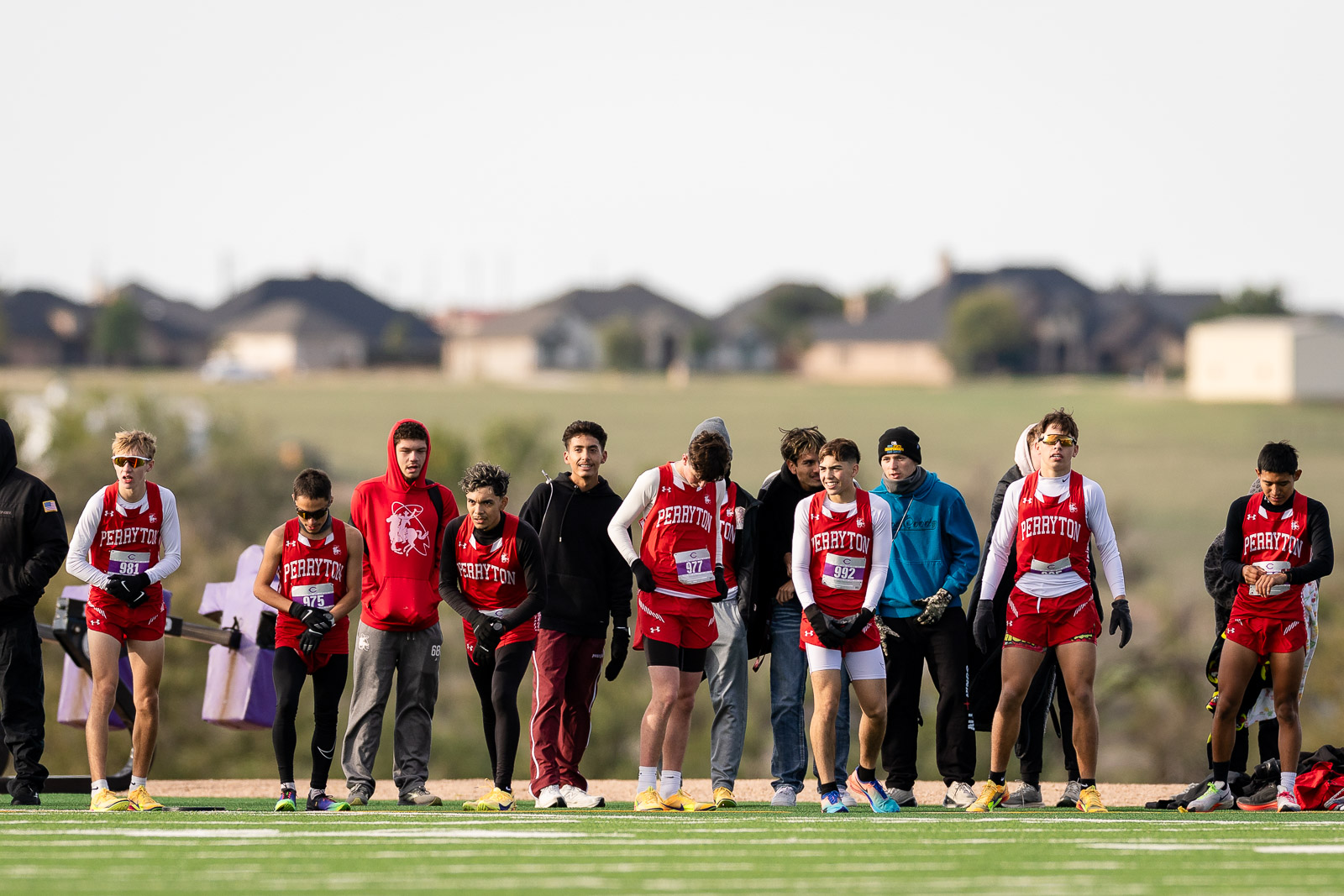 3A-4A-district-xc-jpegJoe-pps-27 | Press Pass Sports Runners in red Perryton uniforms line up at the starting line on a track field, ready to start the race with houses in the background.