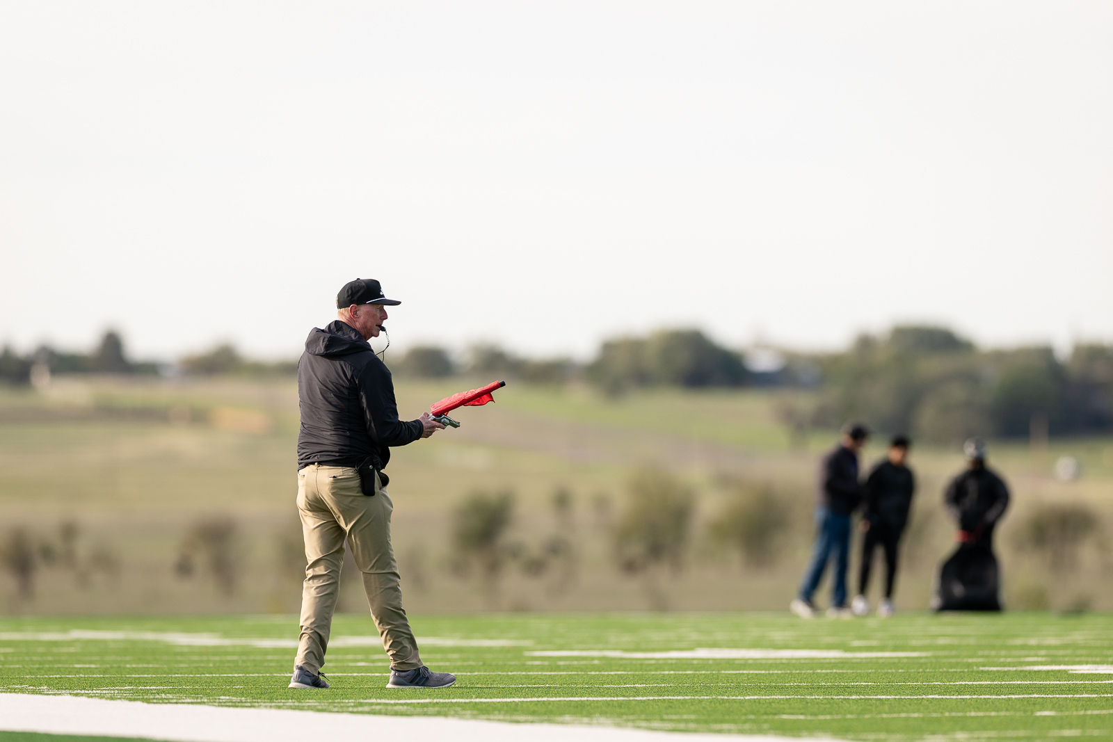 3A-4A-district-xc-jpegJoe-pps-29 | Press Pass Sports Coach on a sports field holding a red flag, wearing a cap and black jacket, with teammates blurred in the background.