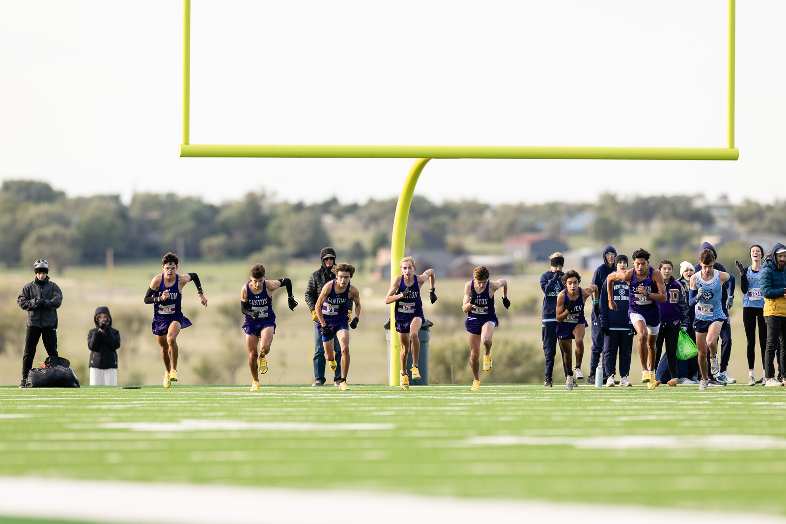 3A-4A-district-xc-jpegJoe-pps-30 | Press Pass Sports Young male sprinters in purple Canyon uniforms race toward the finish line on a bright green track, with coaches and spectators in the background under a yellow goalpost.