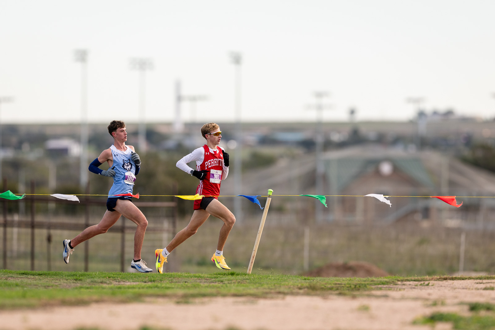 3A-4A-district-xc-jpegJoe-pps-33 | Press Pass Sports Two male runners compete in a cross-country race along a dirt track, one in light blue with a wolf logo and the other in red, with colorful pennant flags in the foreground.