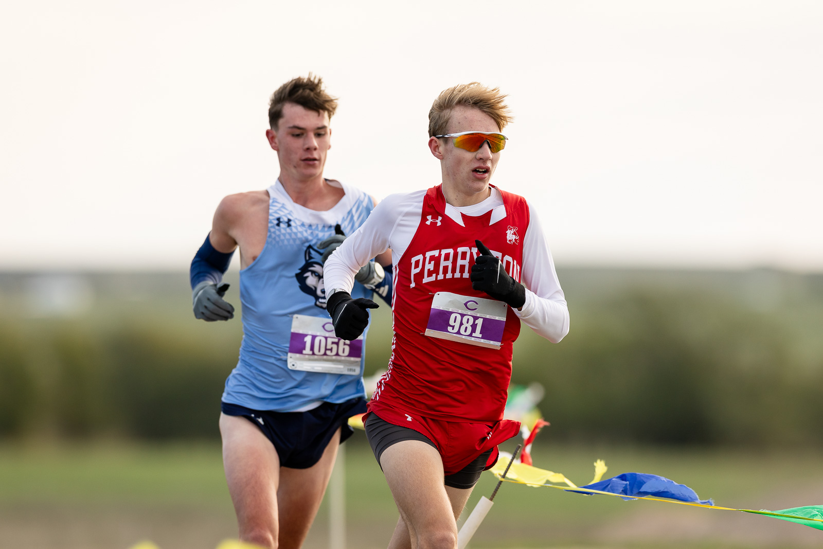 3A-4A-district-xc-jpegJoe-pps-35 | Press Pass Sports Two male runners in a cross-country race; foreground runner in a red jersey with bib 981 and orange sunglasses, behind him a blue jersey with bib 1056.