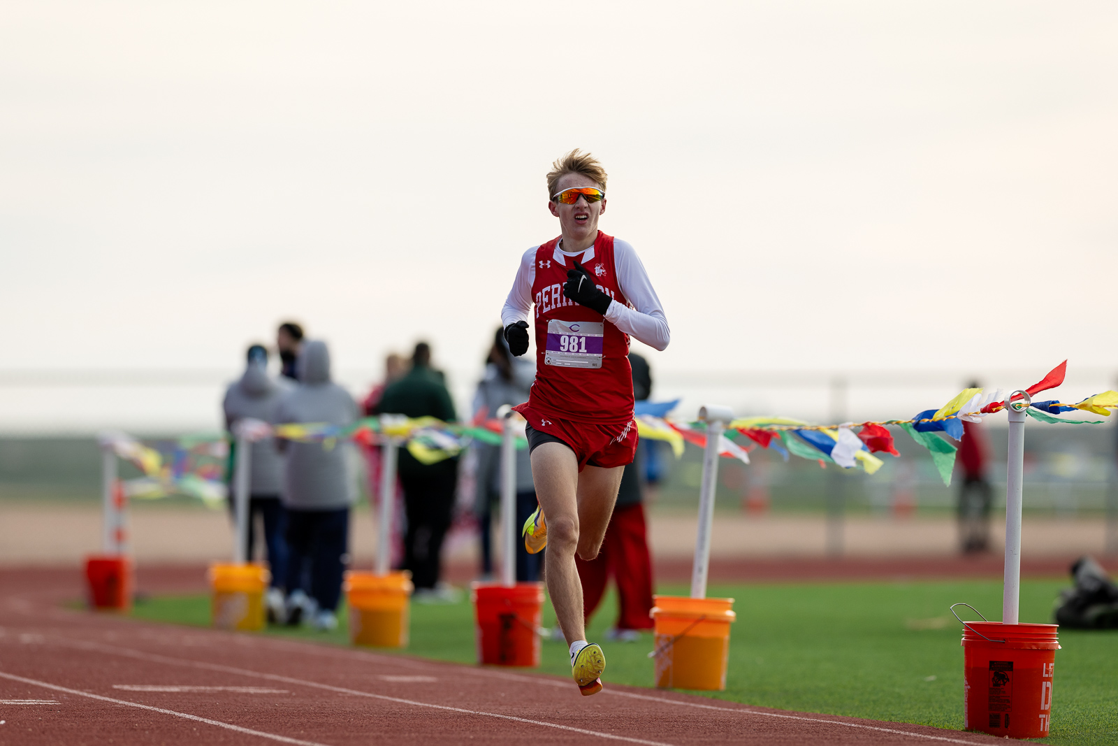 3A-4A-district-xc-jpegJoe-pps-36 | Press Pass Sports Runner in a red uniform crosses the track finish line, bib 981, wearing white sleeves and orange sunglasses. Behind him are blurred spectators and colorful bunting.