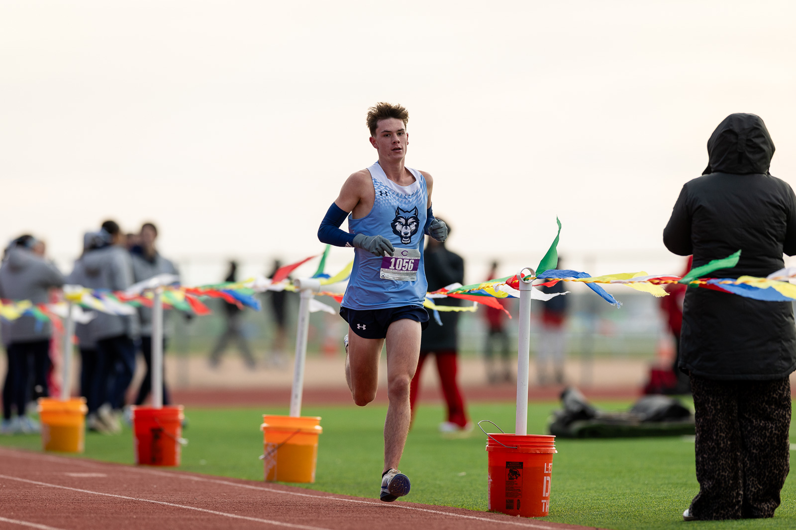 3A-4A-district-xc-jpegJoe-pps-37 | Press Pass Sports A young male runner in a blue jersey crossing the finish line on a track, with spectators and colorful ribbons in the background.