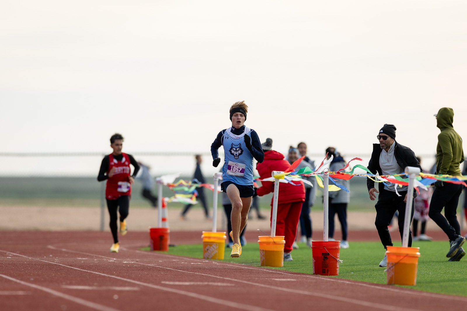 3A-4A-district-xc-jpegJoe-pps-40 | Press Pass Sports Young runner in blue singlet and headband leads a race on a track, finishing near colorful barrier ribbons and orange buckets with spectators in the background.