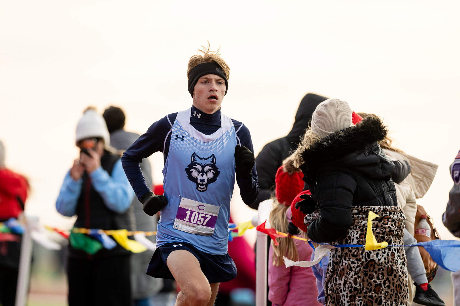 3A-4A-district-xc-jpegJoe-pps-41 | Press Pass Sports Male runner in a blue singlet with a wolf logo, wearing a headband, sprinting during a race with spectators cheering nearby (bib #1057)