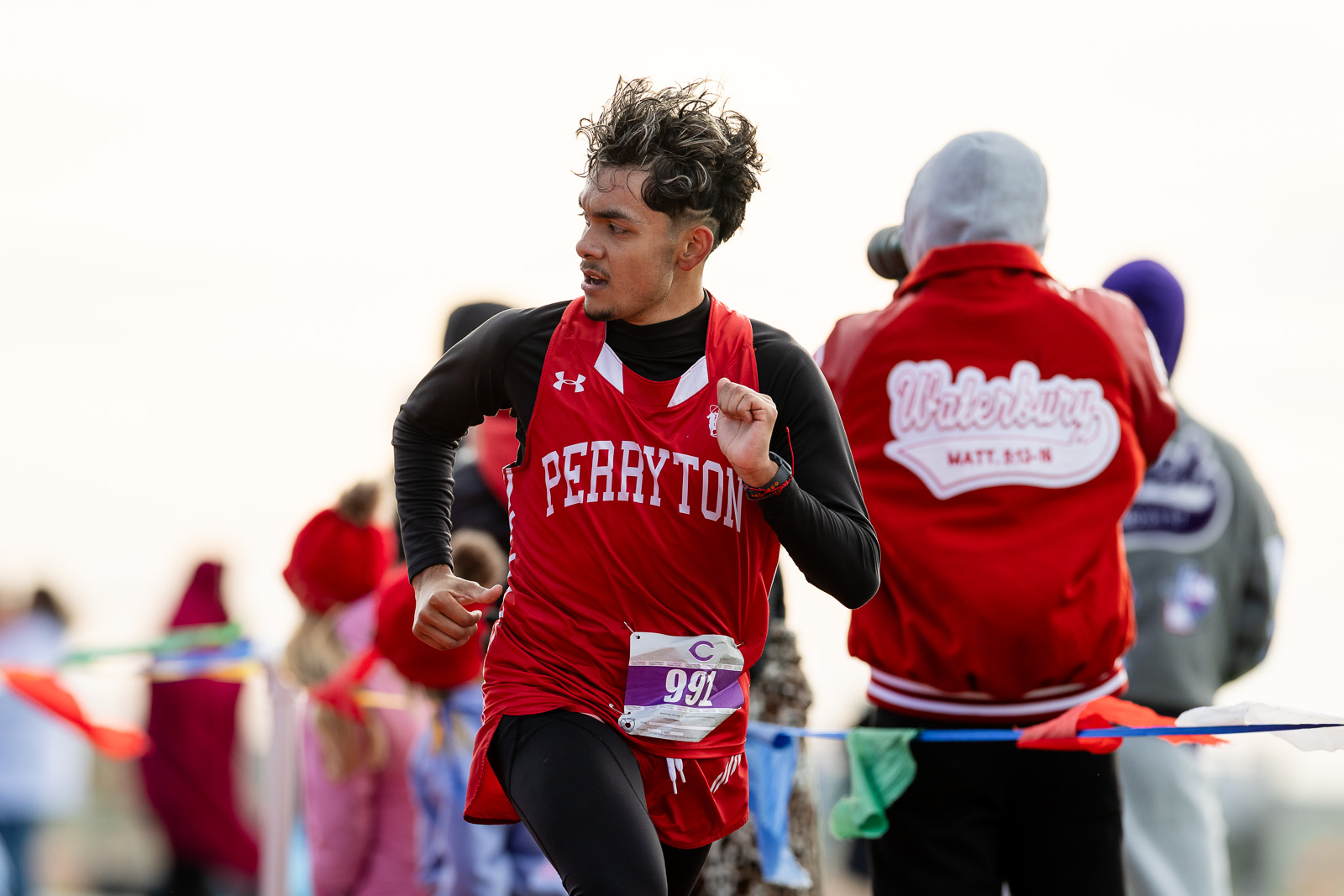 3A-4A-district-xc-jpegJoe-pps-42 | Press Pass Sports Male runner in a red Perryton jersey sprinting in a race, bib number 991, with spectators in the background on a sunny day
