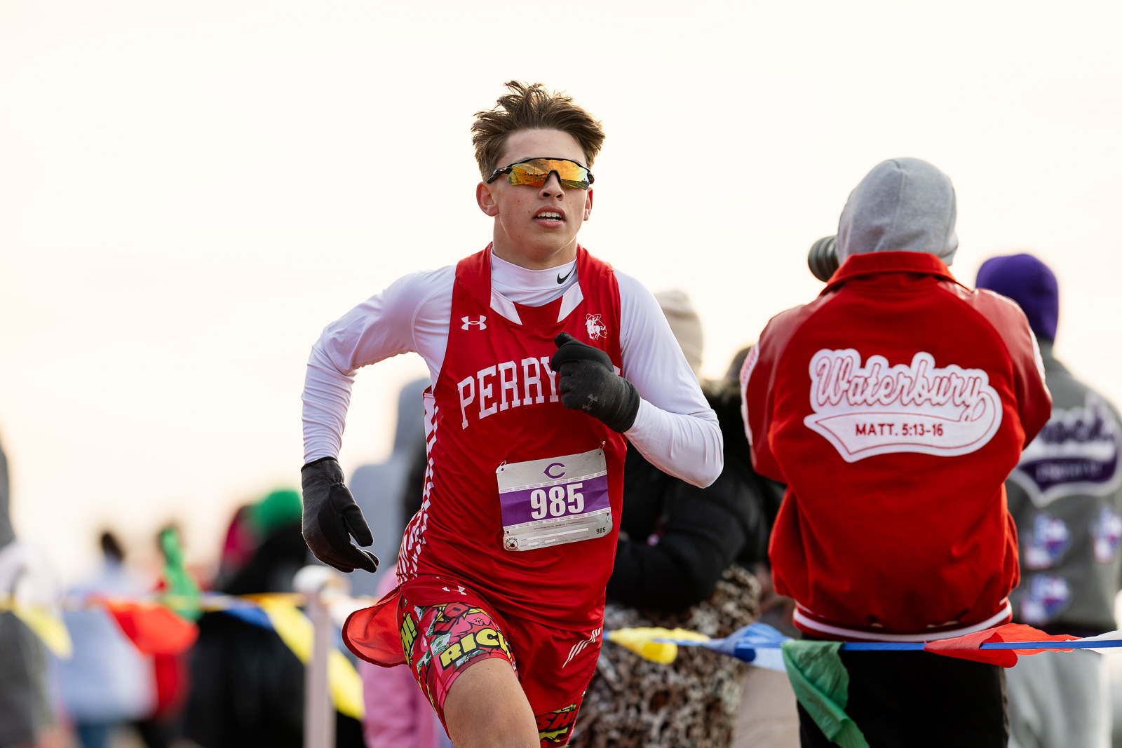 3A-4A-district-xc-jpegJoe-pps-44 | Press Pass Sports Runner in a red Perry team jersey racing forward, bib 985, wearing orange sunglasses and gloves.