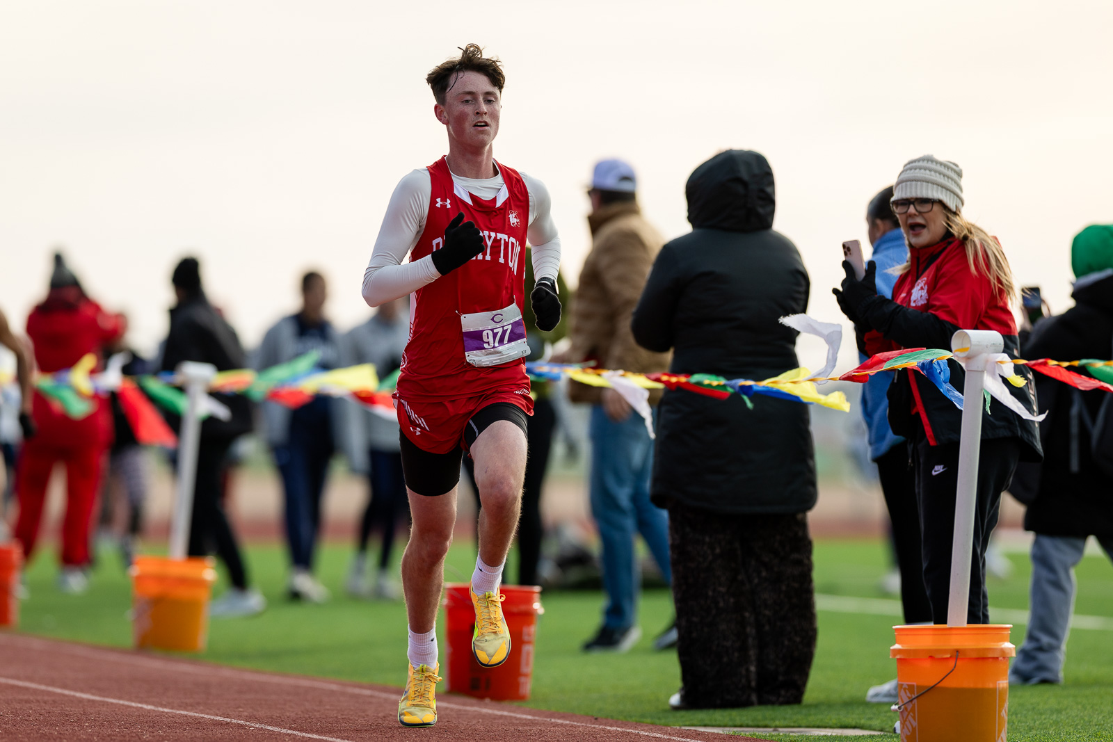 3A-4A-district-xc-jpegJoe-pps-45 | Press Pass Sports Young male runner in red jersey crossing a track finish line, bib 977, with spectators cheering in the background.