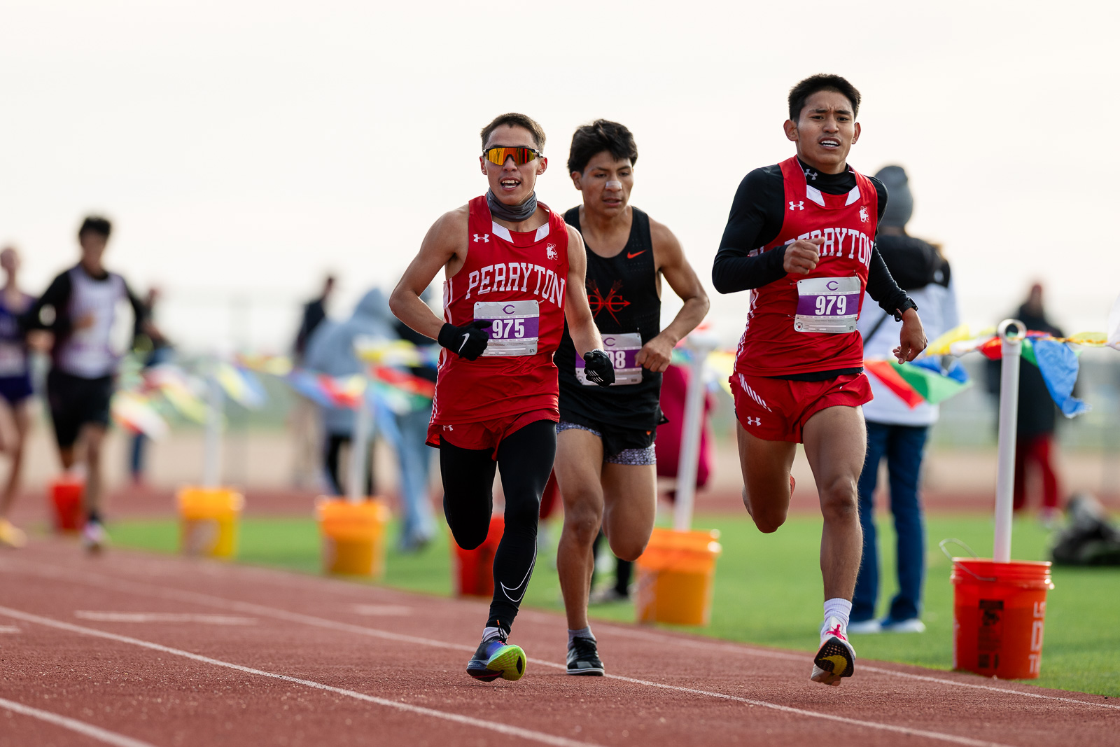 3A-4A-district-xc-jpegJoe-pps-48 | Press Pass Sports Three runners compete on a track; two in red Perryton uniforms lead, one in black behind them, bib numbers visible 975 and 979