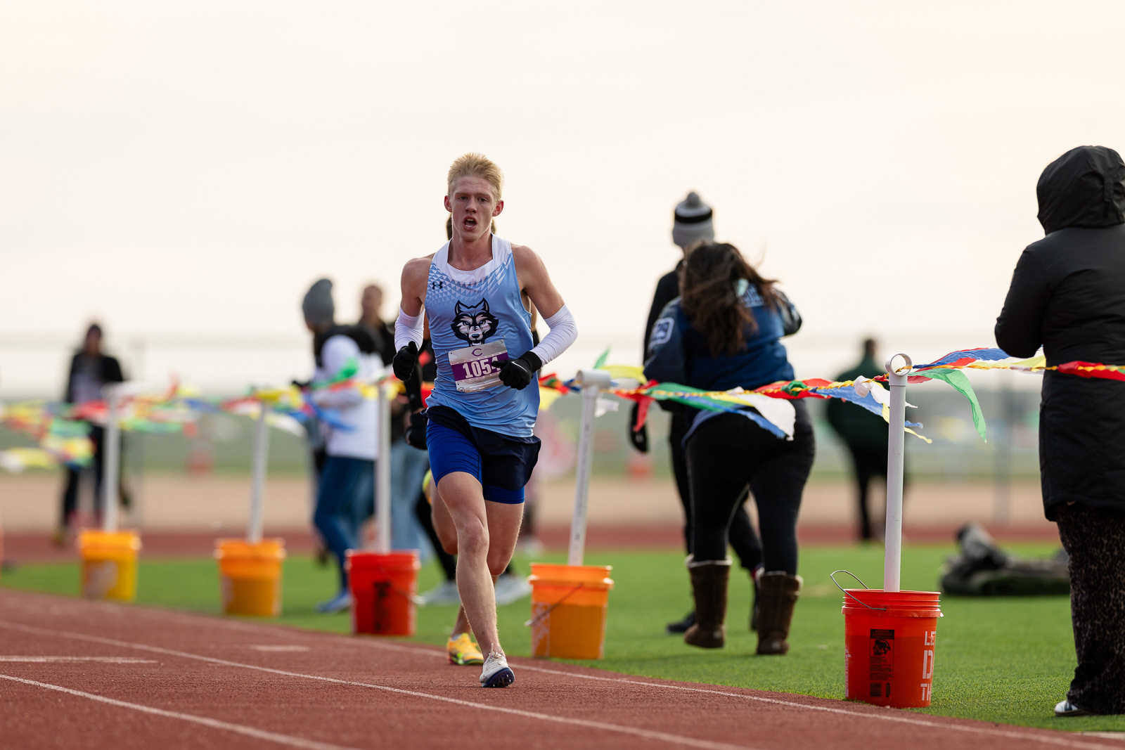 3A-4A-district-xc-jpegJoe-pps-50 | Press Pass Sports Young male runner in a light blue singlet and white arm sleeves nears the finish line on a track, with cheering spectators in the background.