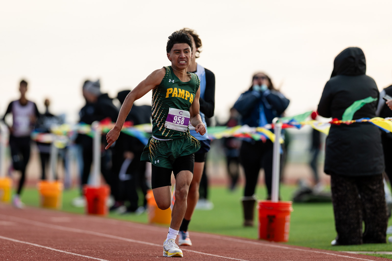 3A-4A-district-xc-jpegJoe-pps-51 | Press Pass Sports Sprinter in a green and yellow uniform with bib 958 runs on a red track while spectators cheer in the background.