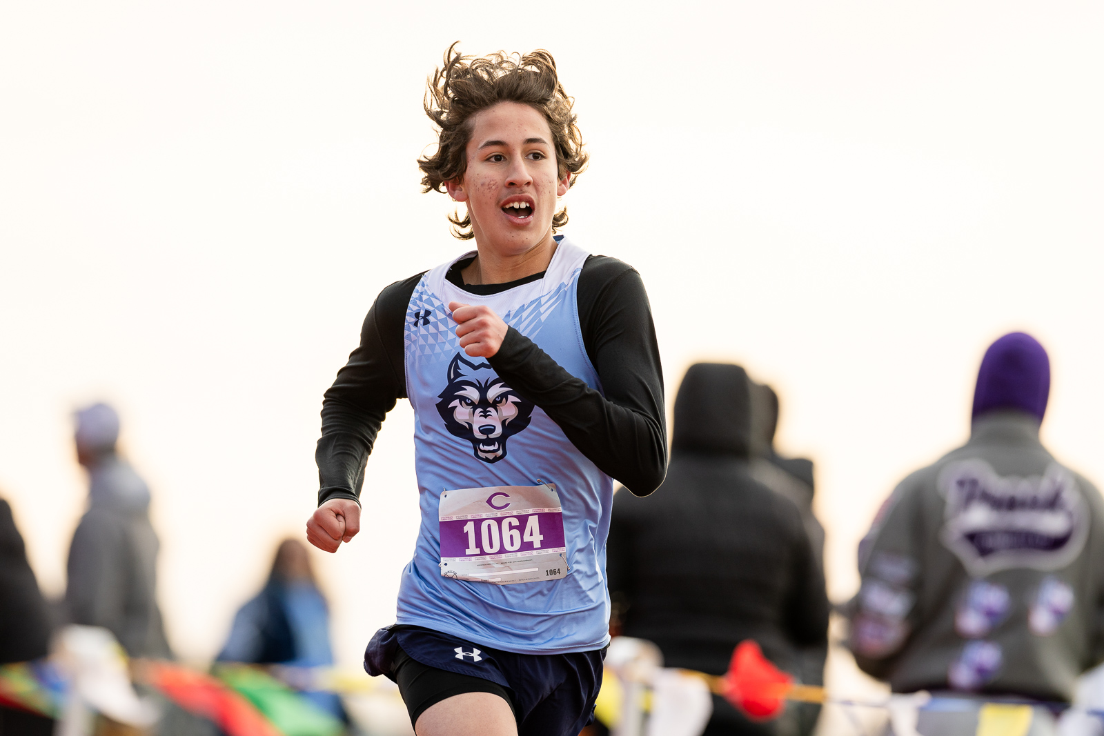 3A-4A-district-xc-jpegJoe-pps-52 | Press Pass Sports Young runner in a blue singlet with a wolf logo, number 1064, sprinting in a race with blurred spectators in the background.