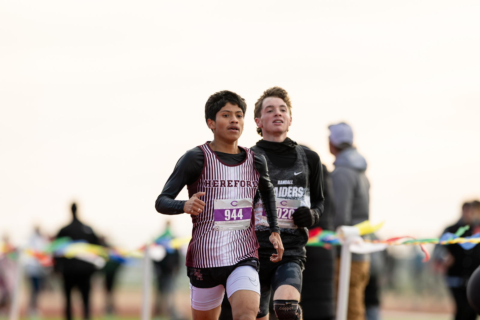 3A-4A-district-xc-jpegJoe-pps-53 | Press Pass Sports Two boys race side by side on a track, bibs 944 and 020, the front runner wearing a maroon-and-white Hereford uniform, with a blurred crowd and colorful finish-line tape in the background.