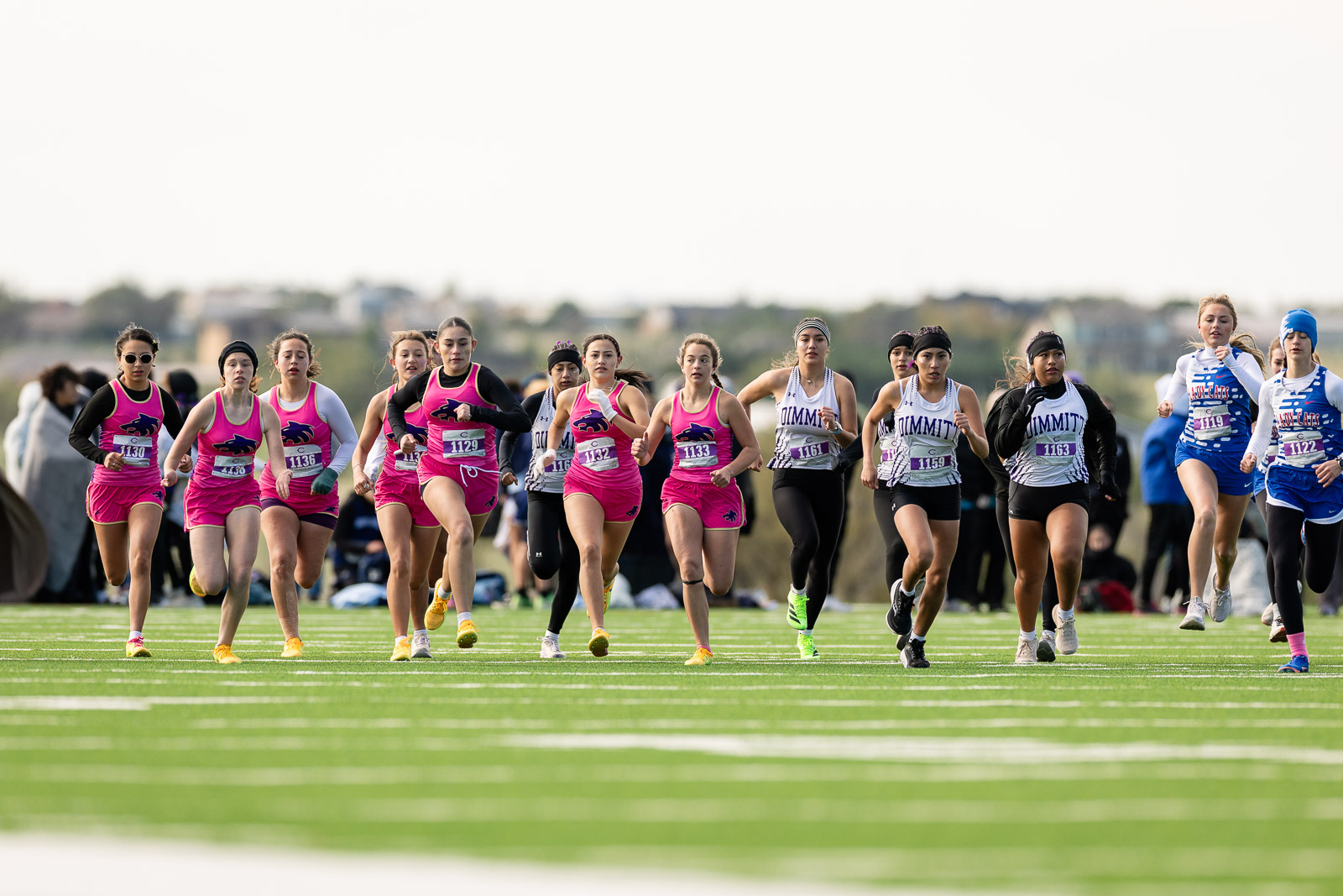 3A-4A-district-xc-jpegJoe-pps-54 | Press Pass Sports Group of female runners at the start of a race on a green track, wearing pink and white uniforms with bib numbers.