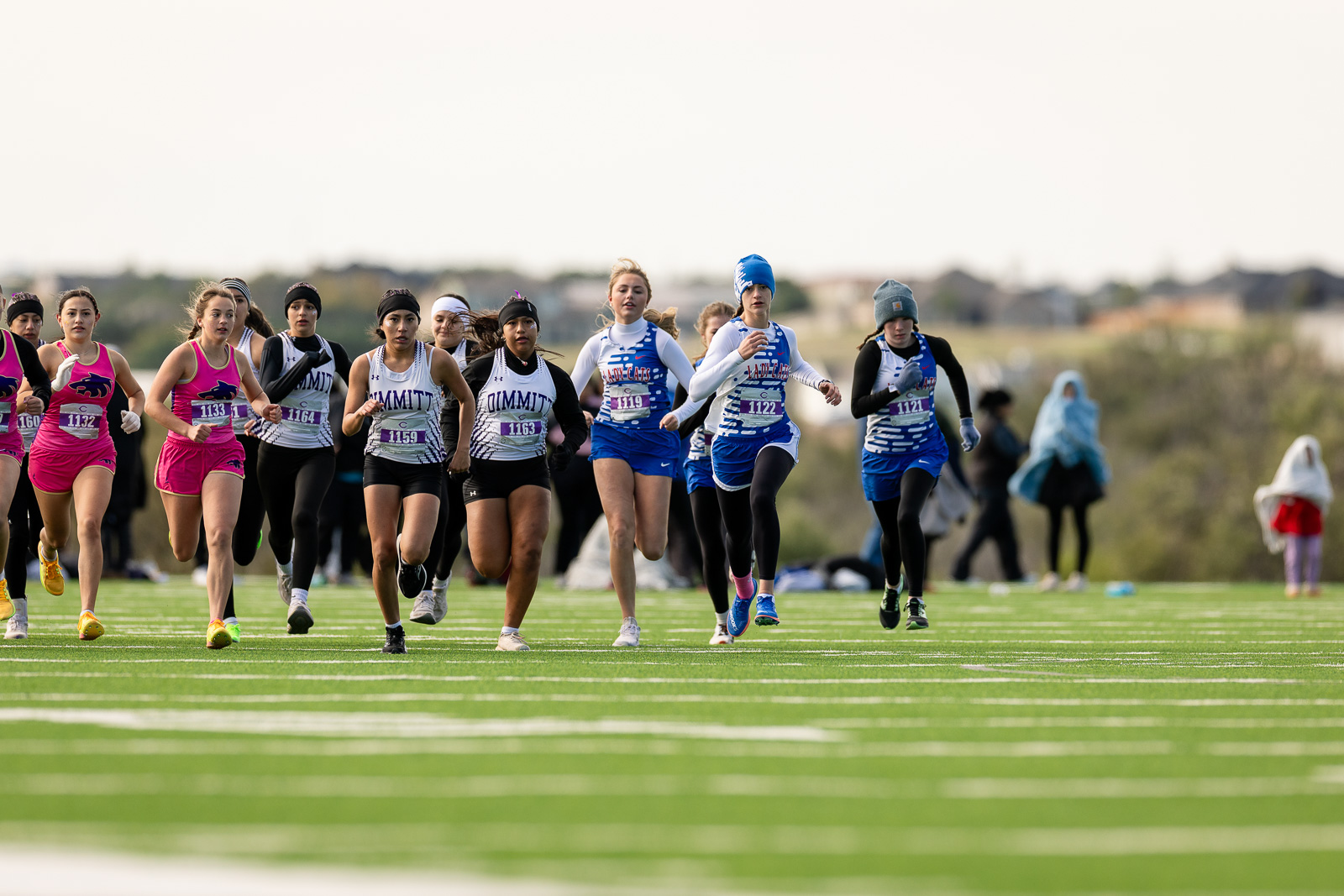 3A-4A-district-xc-jpegJoe-pps-55 | Press Pass Sports Group of female runners at the starting line of a track race, wearing pink and blue uniforms with bib numbers on a green turf field.
