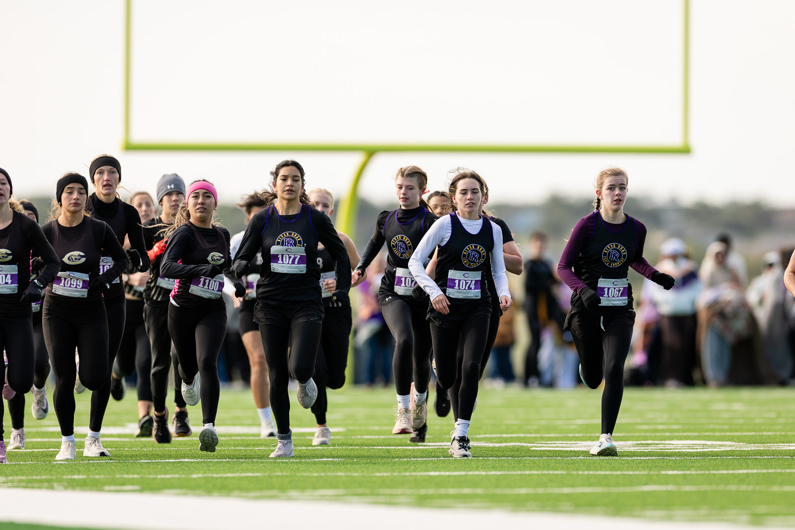 3A-4A-district-xc-jpegJoe-pps-56 | Press Pass Sports Group of female runners at the start of a race on a football field, wearing black uniforms and numbered bibs.