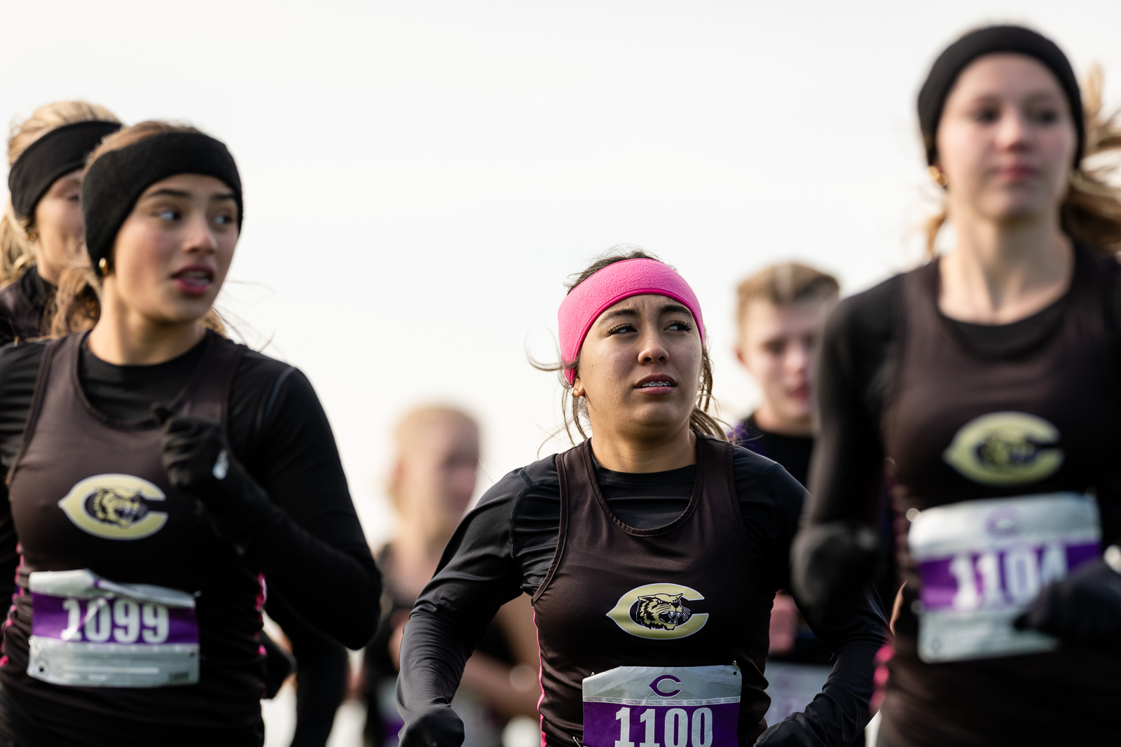 3A-4A-district-xc-jpegJoe-pps-59 | Press Pass Sports Group of runners in black athletic outfits racing, main focus on a woman with a pink headband wearing bib 1100.
