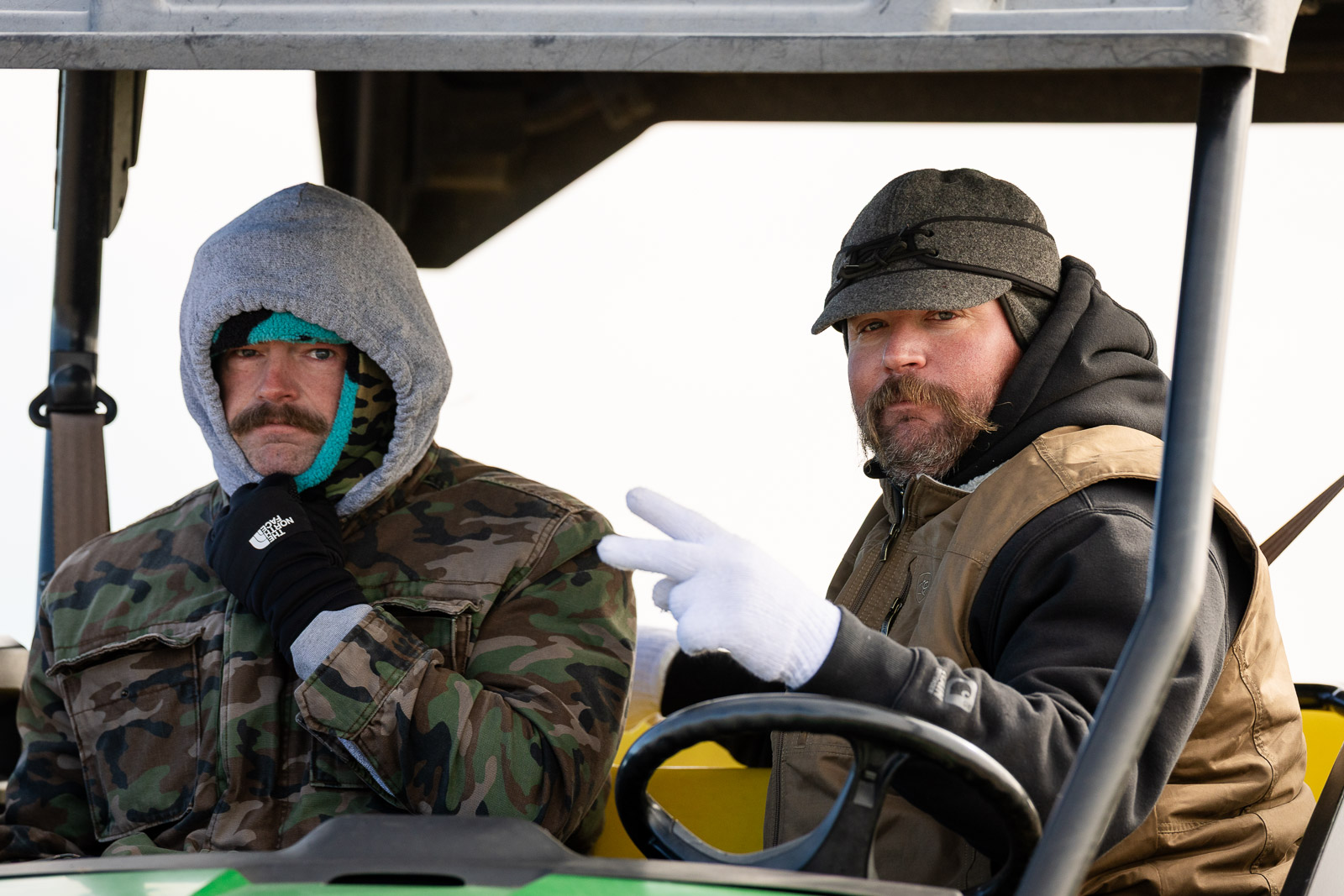 3A-4A-district-xc-jpegJoe-pps-60 | Press Pass Sports Two men in a cab wearing warm jackets; one in camouflage with a gray hood, the other in a cap and brown vest, looking at the camera.