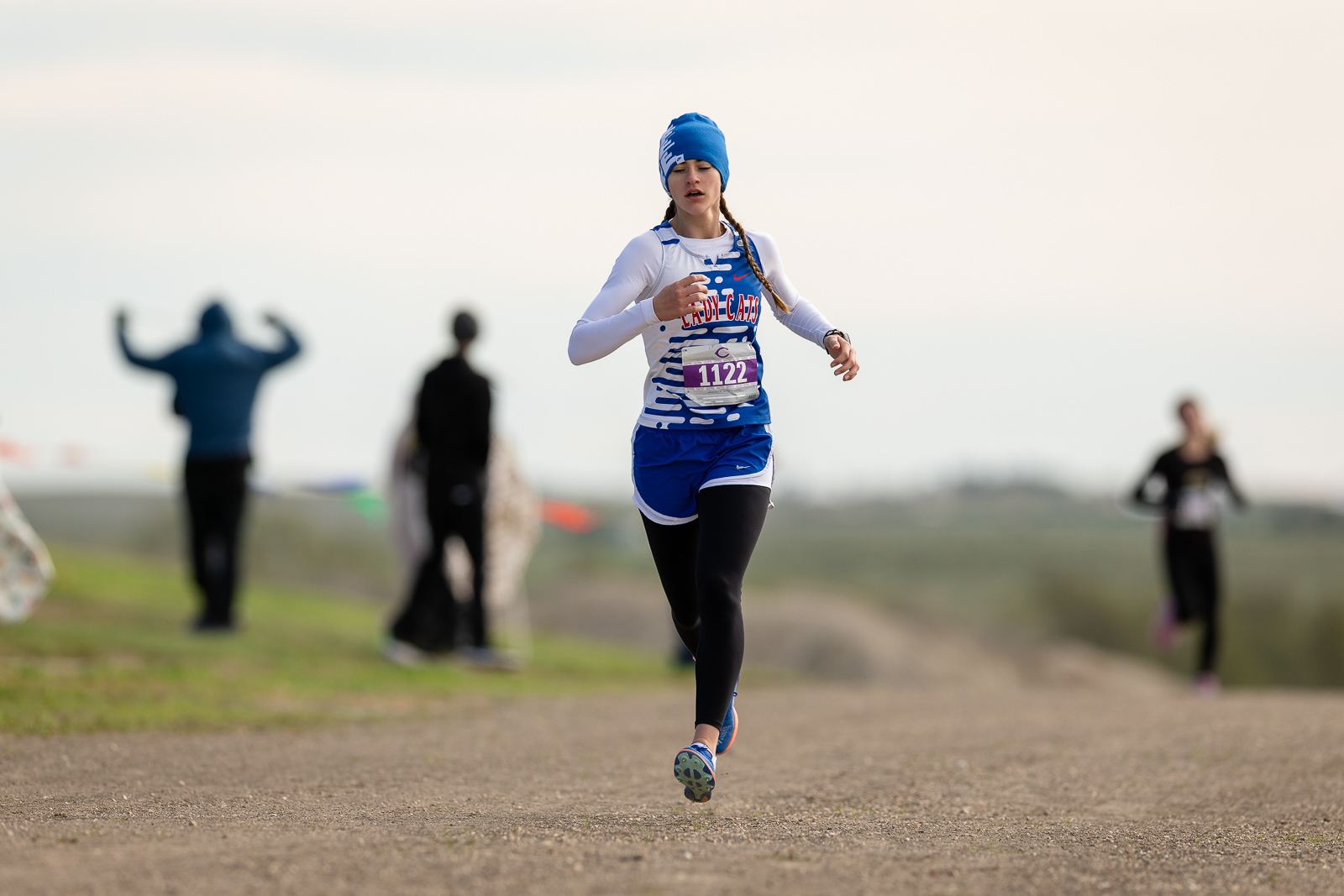 3A-4A-district-xc-jpegJoe-pps-61 | Press Pass Sports Female runner in blue and white outfit, bib 1122, sprinting on a dirt path with spectators blurred in the background.