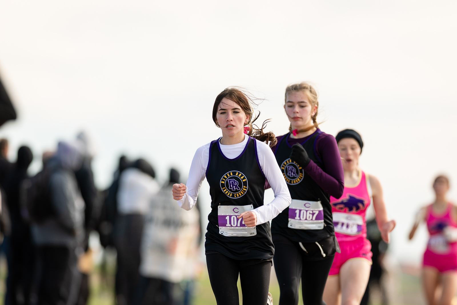 3A-4A-district-xc-jpegJoe-pps-63 | Press Pass Sports Female runner in a black and purple jersey leading a race, bib 1074, with spectators blurred in the background on a sunny day.