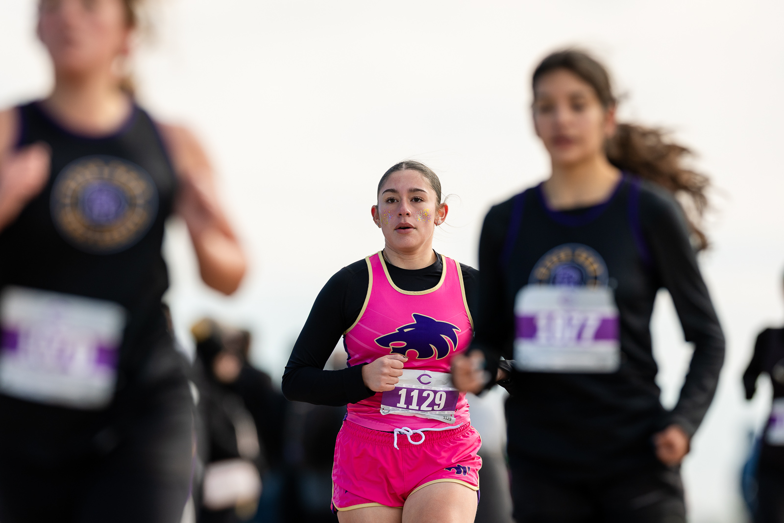 3A-4A-district-xc-jpegJoe-pps-66 | Press Pass Sports Young female runner in a bright pink athletic outfit with glitter on her cheeks, bib number 1129, mid-race among blurred runners.