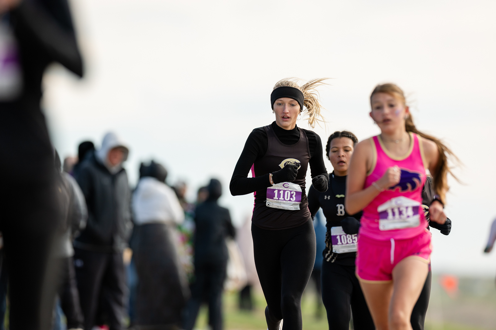 3A-4A-district-xc-jpegJoe-pps-67 | Press Pass Sports Female runner in black with a headband, bib 1103, sprinting toward the camera with spectators on the side of the course.