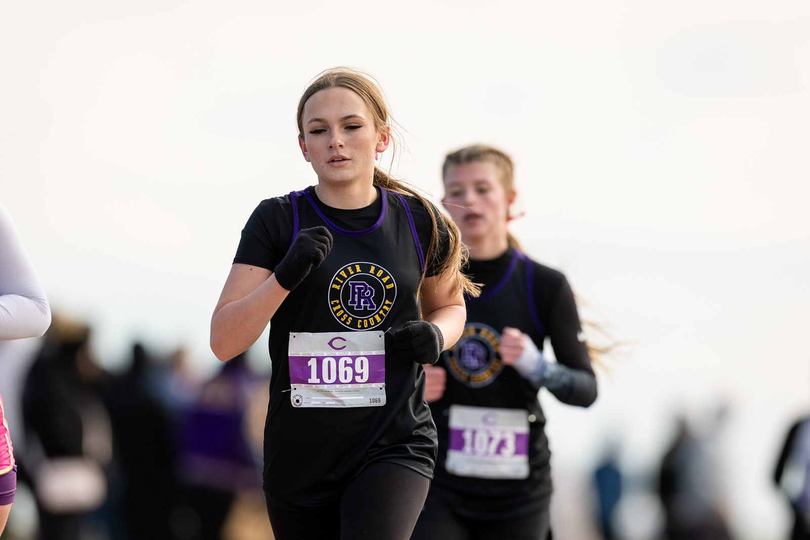 3A-4A-district-xc-jpegJoe-pps-69 | Press Pass Sports Young woman running in a cross-country race, wearing a black shirt with purple trim and gloves, bib number 1069, focused expression.