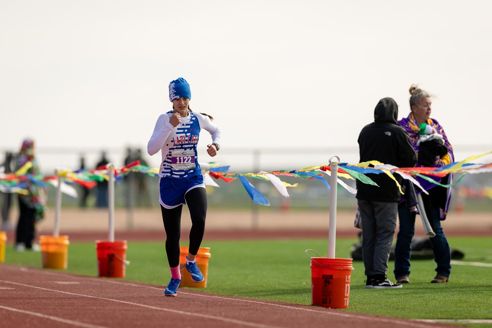 3A-4A-district-xc-jpegJoe-pps-71 | Press Pass Sports Female runner in a blue-and-white outfit and blue beanie sprinting on a track toward the finish line, bib number 1122, with colorful pennant tape nearby.