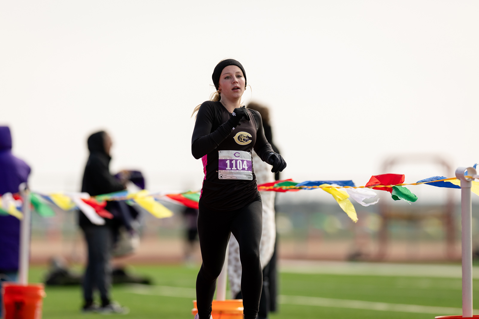 3A-4A-district-xc-jpegJoe-pps-72 | Press Pass Sports Female runner in black athletic wear and headband, bib 1104, mid-race on a track with colorful banners in the foreground.