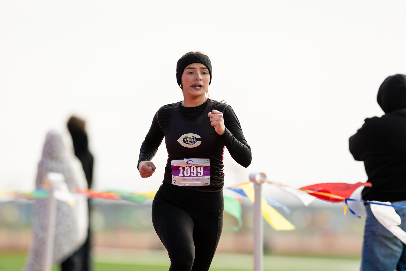 3A-4A-district-xc-jpegJoe-pps-73 | Press Pass Sports Female runner in a black outfit wearing a headband, mid-stride with bib 1099 during a race, colorful flags in the background.