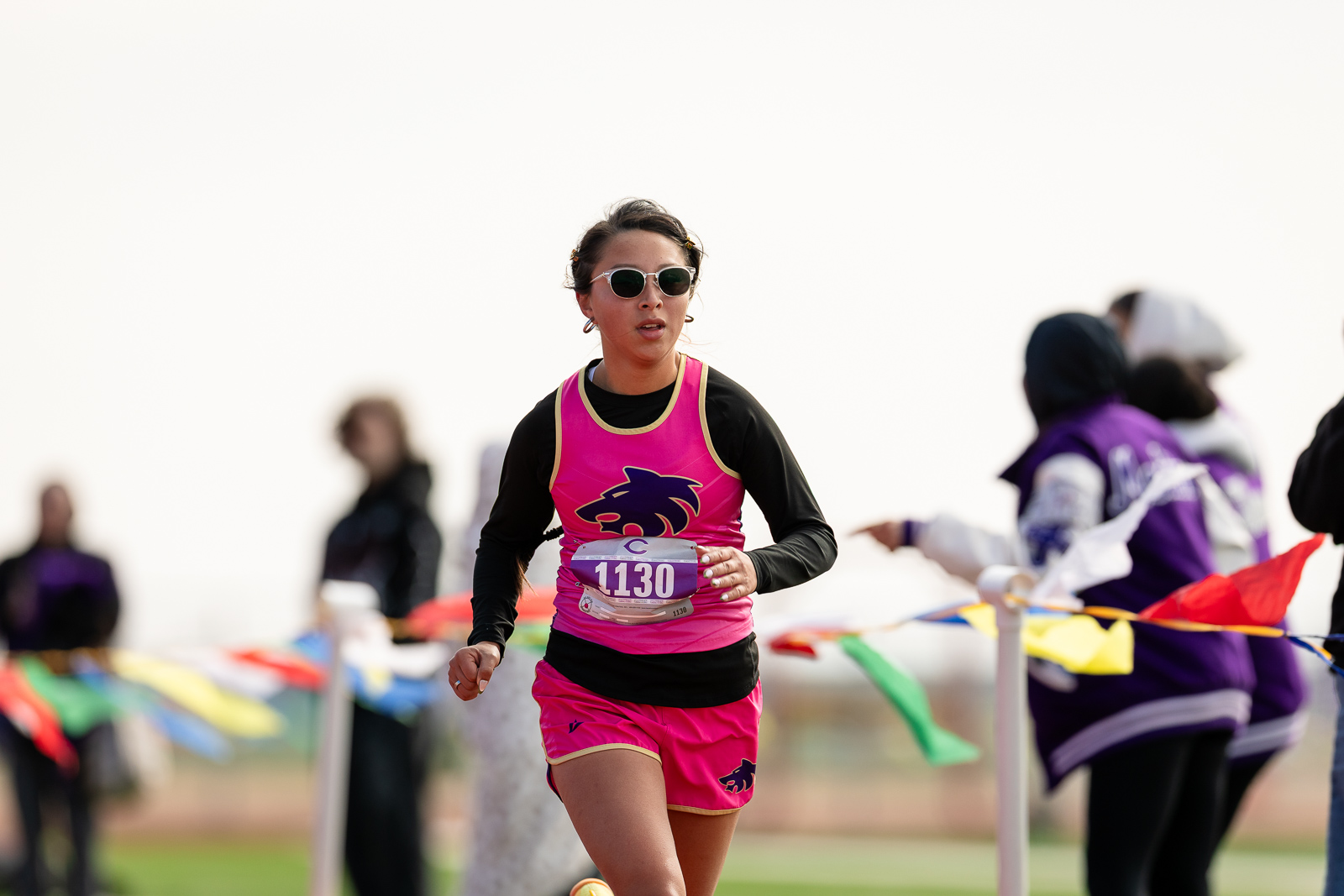 3A-4A-district-xc-jpegJoe-pps-75 | Press Pass Sports Female runner in pink and black athletic wear, bib 1130, running in a race with spectators in the background, wearing sunglasses.