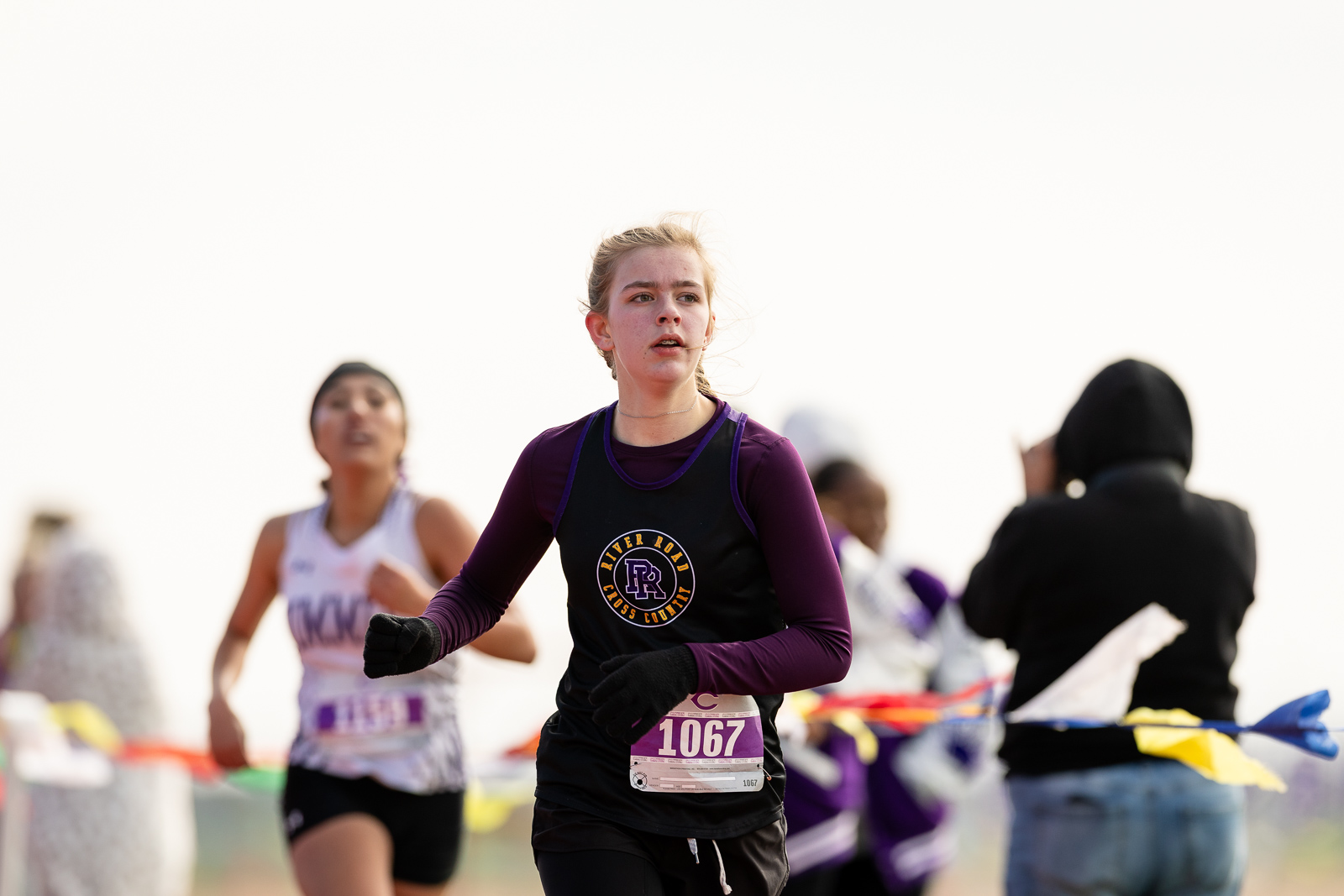 3A-4A-district-xc-jpegJoe-pps-76 | Press Pass Sports Front-running female runner in a maroon long-sleeve and black singlet with bib 1067 during a cross-country race, others blurred in the background.