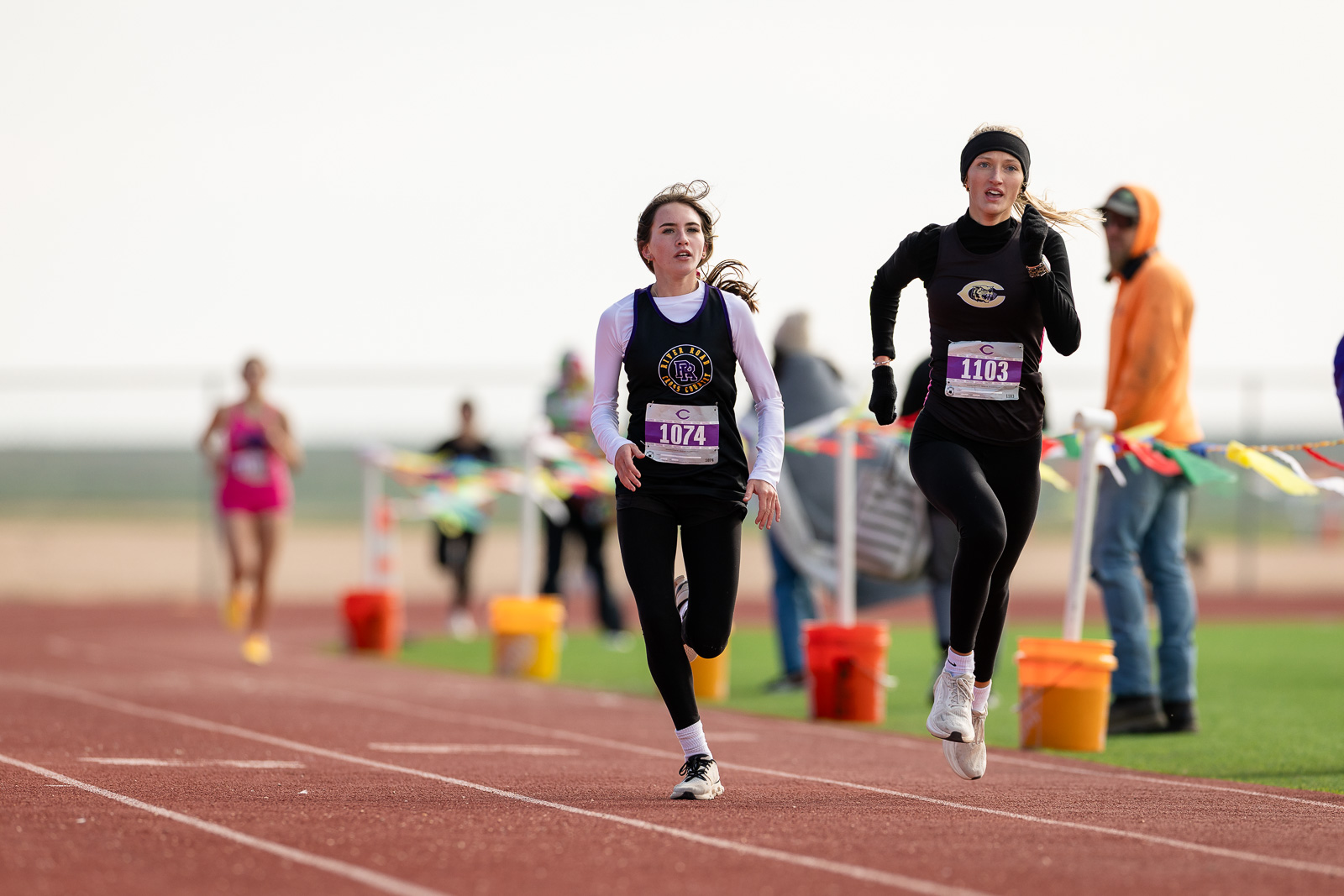 3A-4A-district-xc-jpegJoe-pps-78 | Press Pass Sports Two female runners sprint toward the finish on a track, wearing race bibs 1074 and 1103, with spectators in the background.