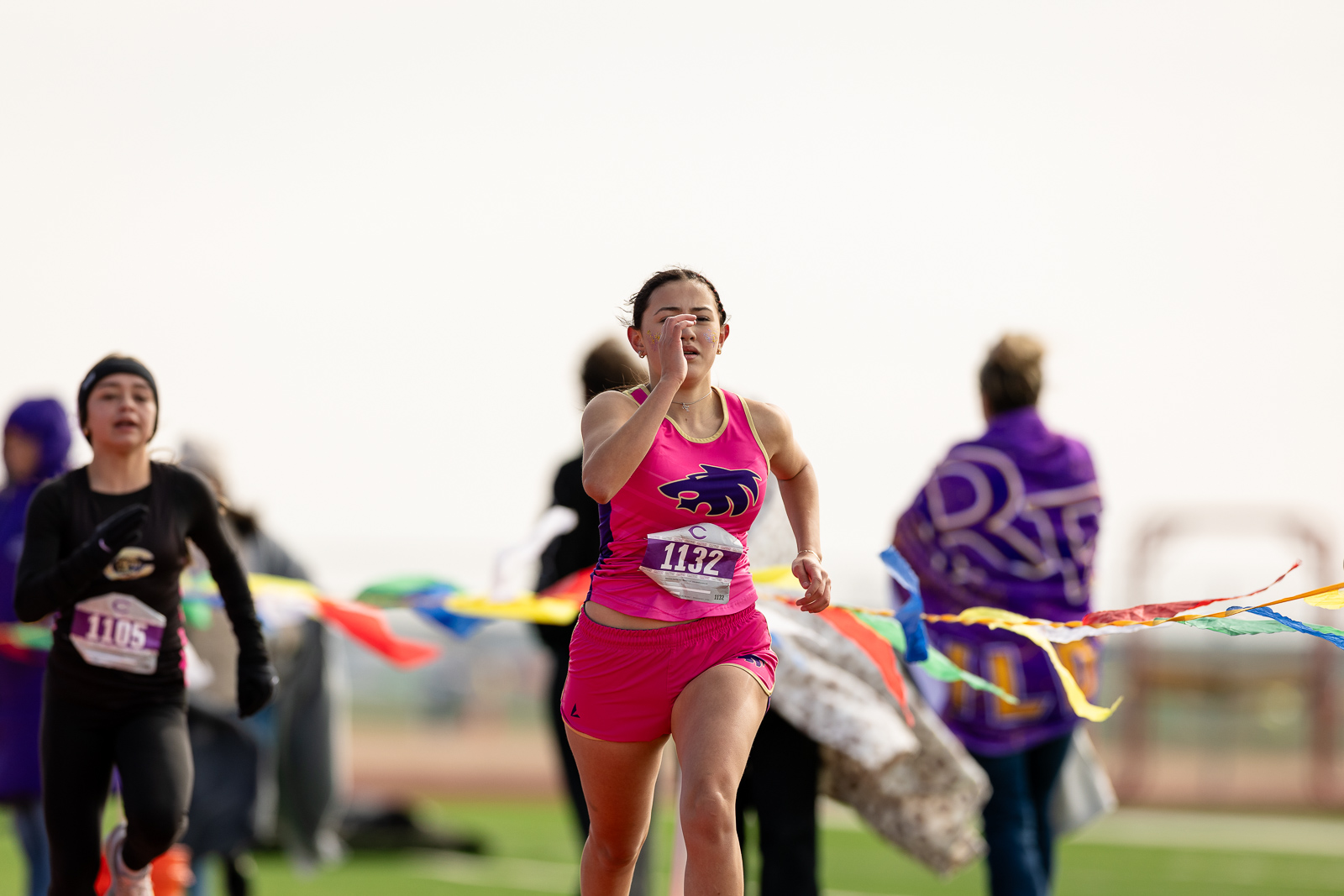 3A-4A-district-xc-jpegJoe-pps-80 | Press Pass Sports Young female runner in bright pink athletic kit (bib 1132) sprinting toward the finish line on a track with colorful ribbons in the foreground.