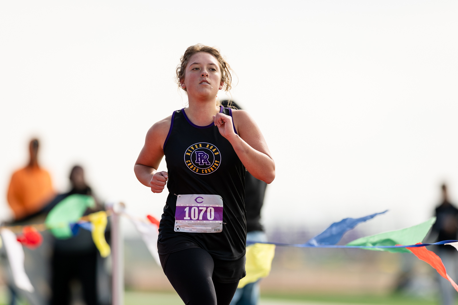 3A-4A-district-xc-jpegJoe-pps-82 | Press Pass Sports Female cross-country runner in a black tank top with bib 1070, running with determination among colorful course bunting.