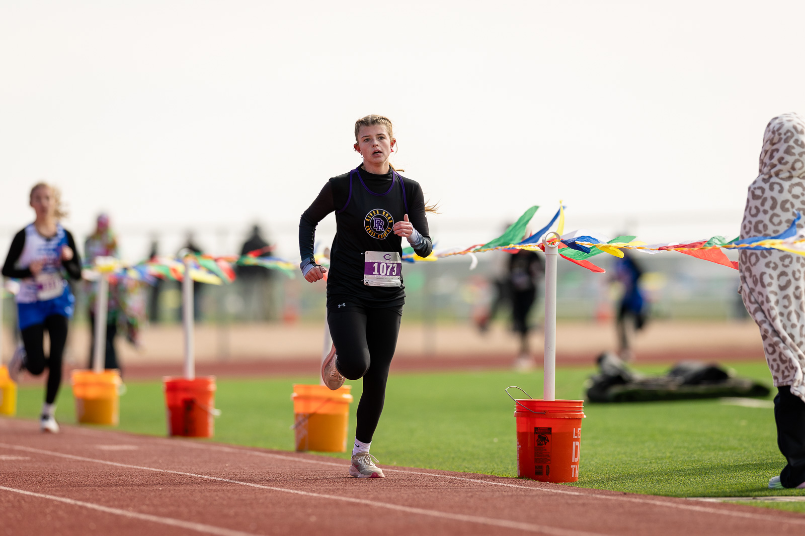 3A-4A-district-xc-jpegJoe-pps-83 | Press Pass Sports Female runner in black athletic wear with bib 1073 sprinting on a track toward the finish line with colorful ribbons in the background.