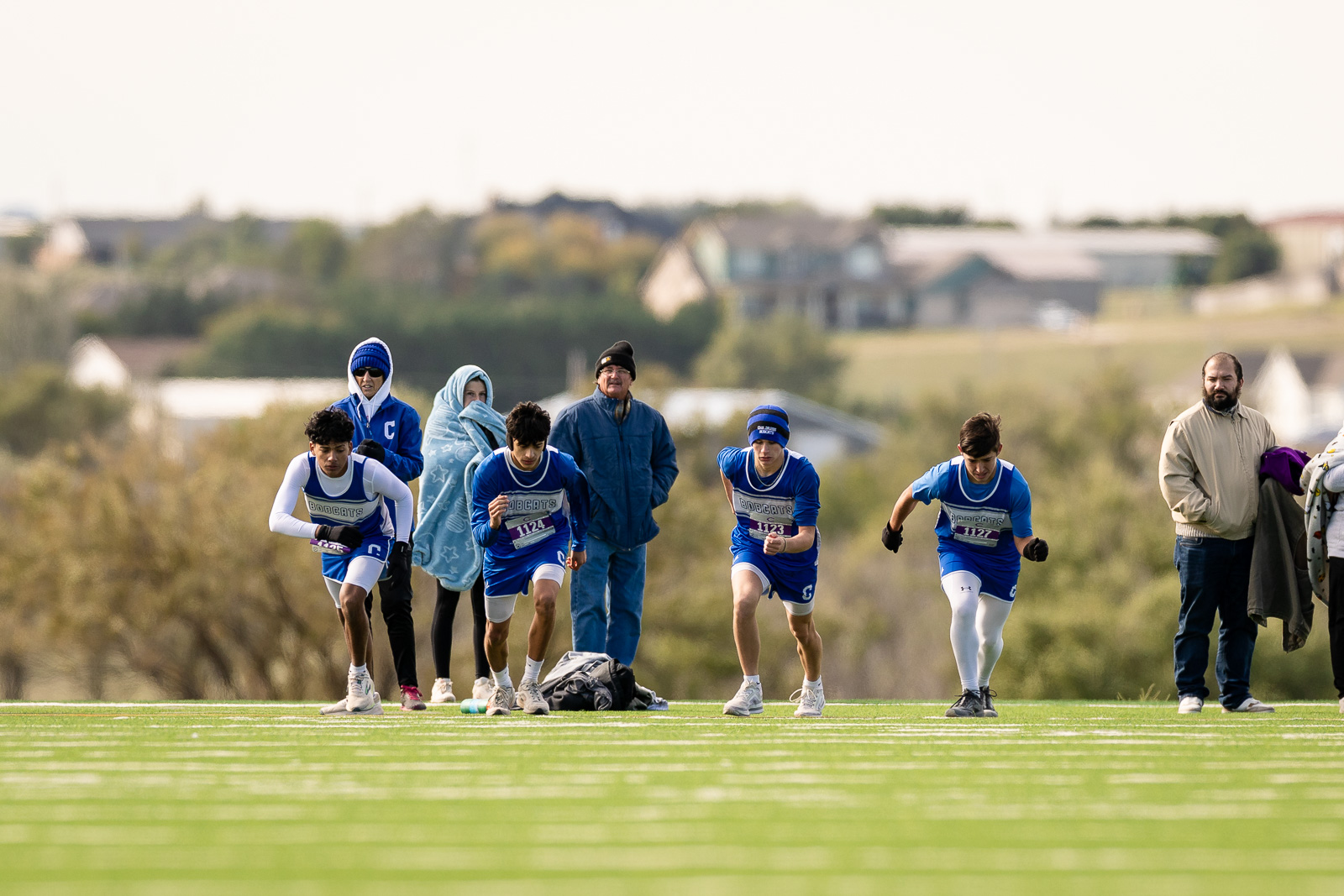 3A-4A-district-xc-jpegJoe-pps-85 | Press Pass Sports Young athletes in blue uniforms crouch at the starting line on a grassy track, awaiting the race start, with spectators in the background.