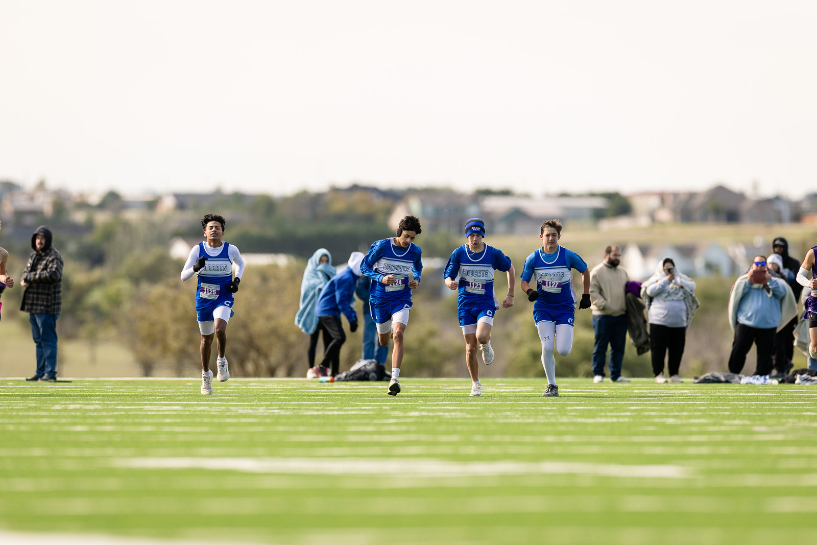 3A-4A-district-xc-jpegJoe-pps-86 | Press Pass Sports Four male runners in blue uniforms sprint along a green track, with spectators watching from the sideline.