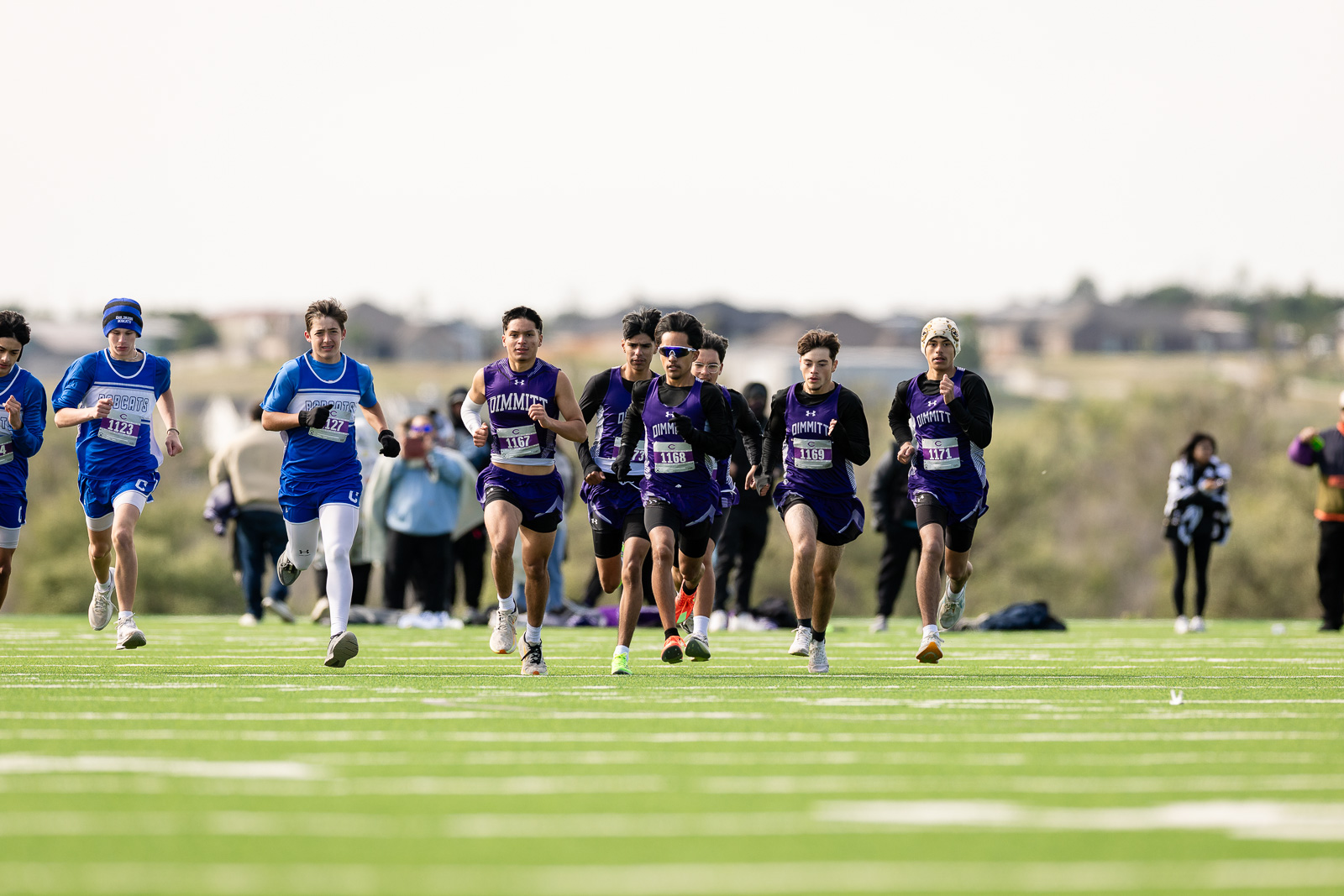 3A-4A-district-xc-jpegJoe-pps-87 | Press Pass Sports Group of runners in blue and purple uniforms sprint on a green track, with spectators and a blurred background.
