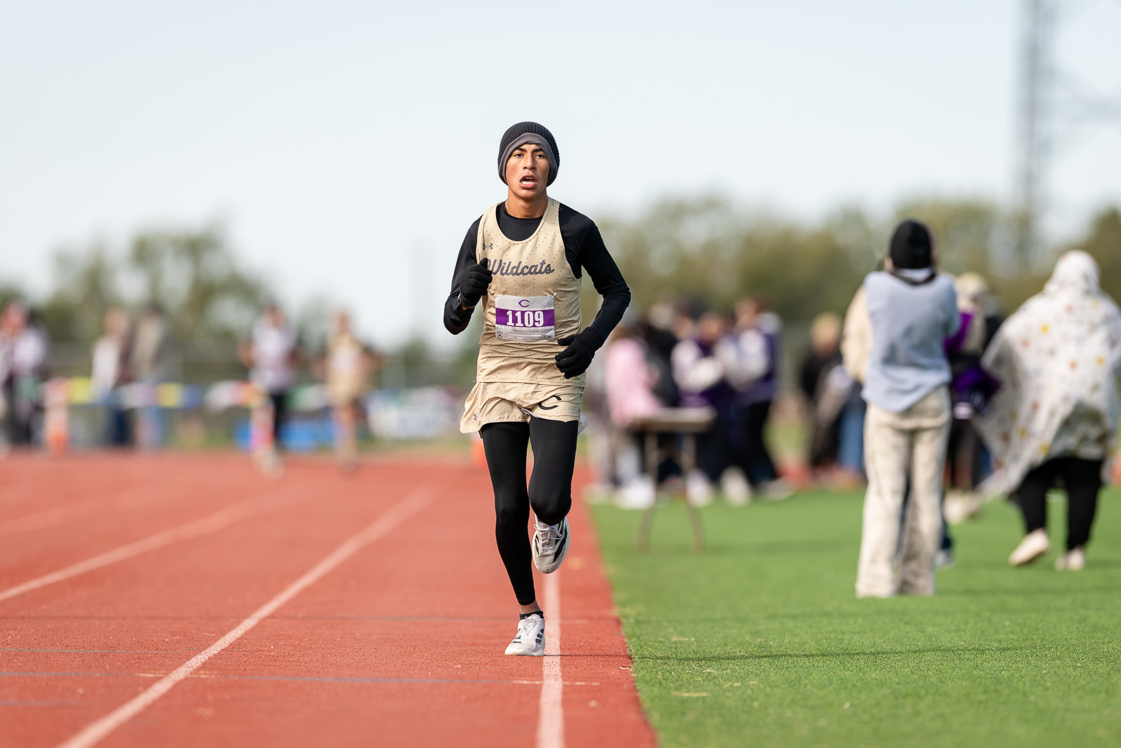 3A-4A-district-xc-jpegJoe-pps-91 | Press Pass Sports Young male runner in a beige racing kit and black sleeves, wearing a beanie, sprinting on a red track with bib number 1109 while blurred spectators watch in the background.
