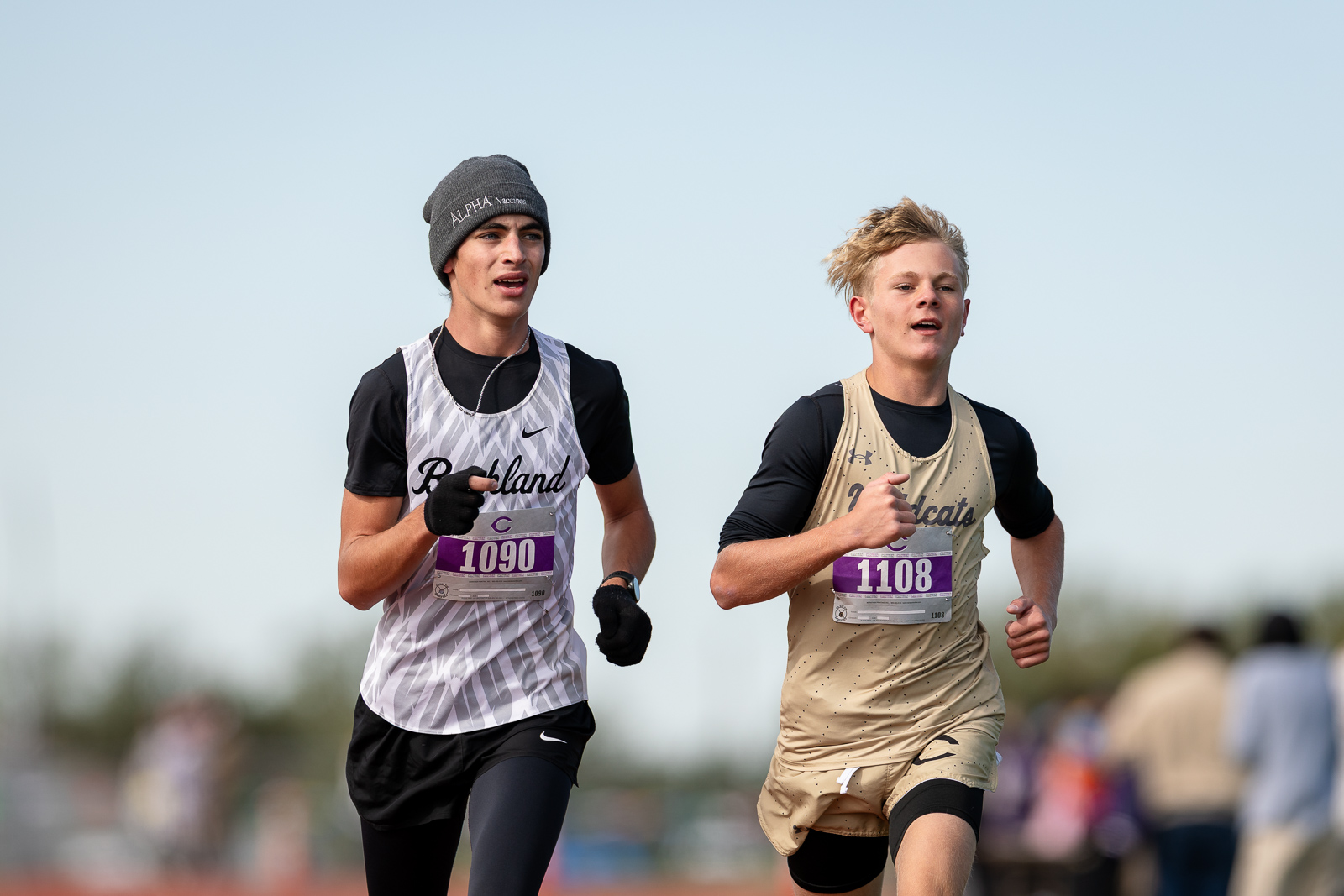 3A-4A-district-xc-jpegJoe-pps-92 | Press Pass Sports Two young male runners in a race, wearing numbered bibs (1090 and 1108) and athletic gear, sprinting side by side outdoors under a clear sky.