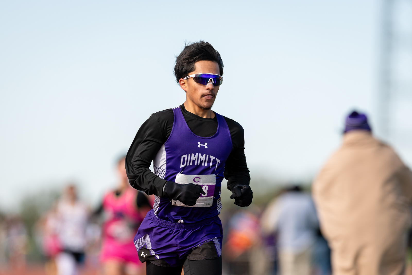 3A-4A-district-xc-jpegJoe-pps-93 | Press Pass Sports Male runner in a purple Dimmitt/Dimmittt singlet with black sleeves, wearing sporty sunglasses, sprinting in a daytime road race with blurred spectators in the background.