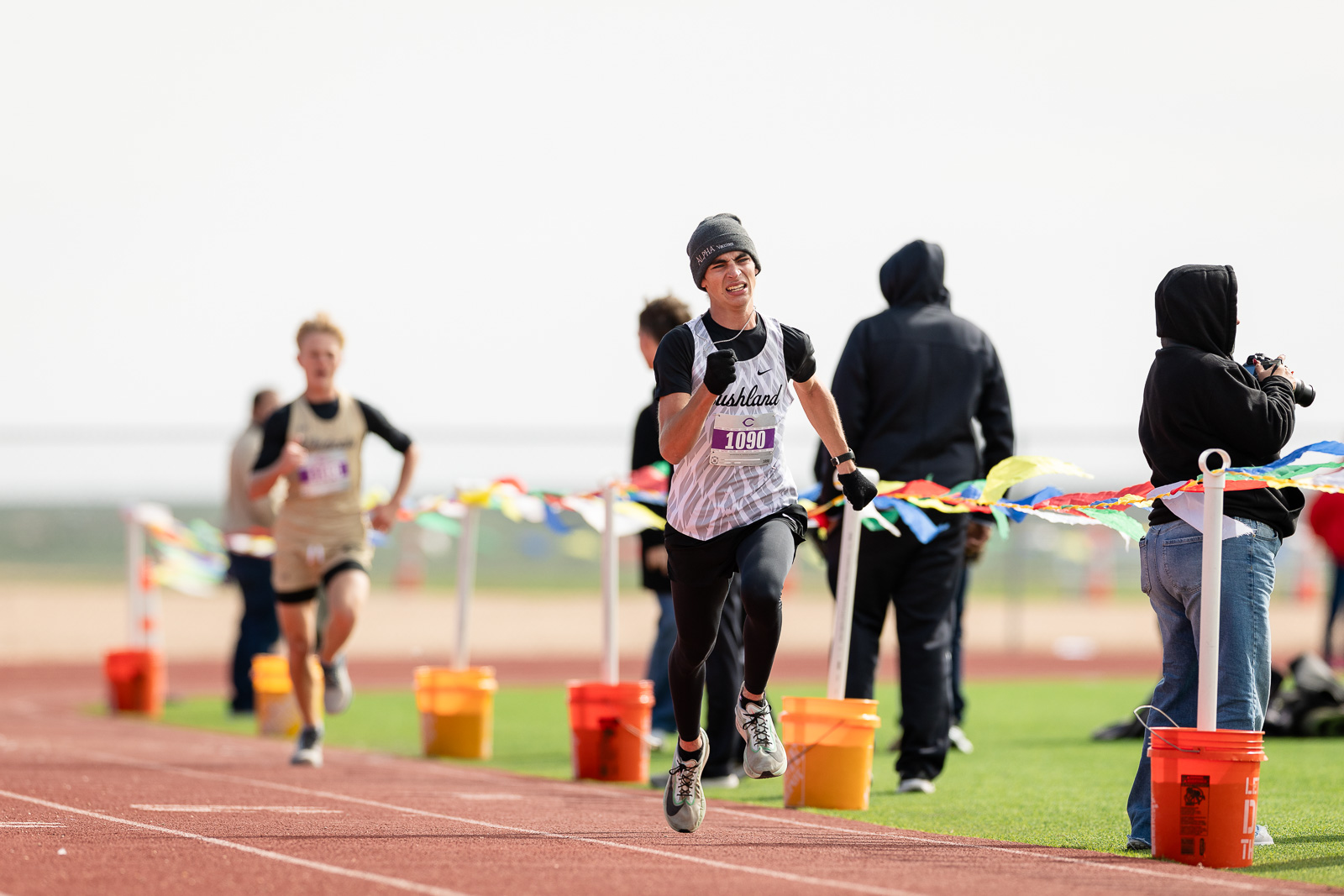 3A-4A-district-xc-jpegJoe-pps-96 | Press Pass Sports Male sprinter in a white sleeveless jersey with bib 1090, running on a red track toward the finish line as colorful pennants flutter nearby.